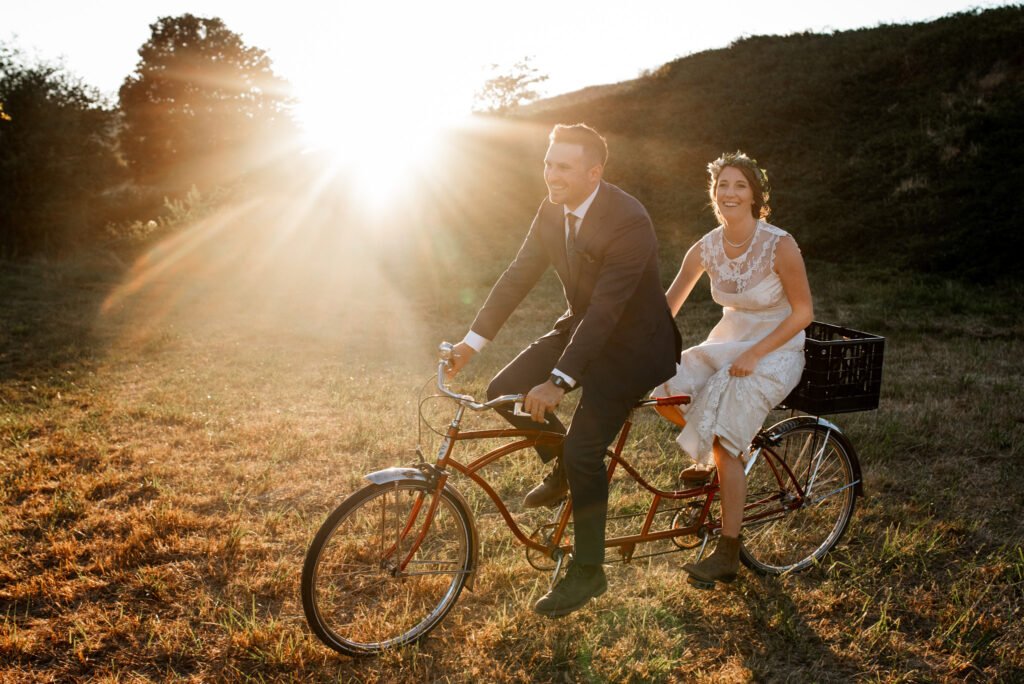 A bride and groom ride a tandem bike at sunset, a perfect elopement activity.