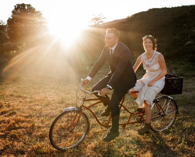 A bride and groom ride a tandem bike at sunset, a perfect elopement activity.