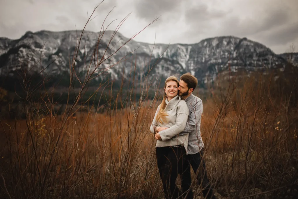A couple embraces near Beacon Rock State Park with snow-capped mountains as a backdrop. With cloudy skies setting a serene atmosphere, this moment is captured perfectly by a Portland wedding and elopement photographer.