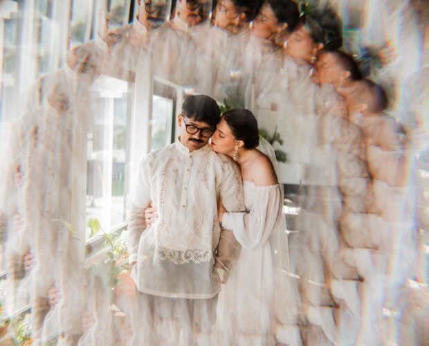 A couple in traditional wedding attire stands by a window with various plants, surrounded by a burst of colors. They are celebrating their wedding at Union/Pine, blending classic and modern elements.