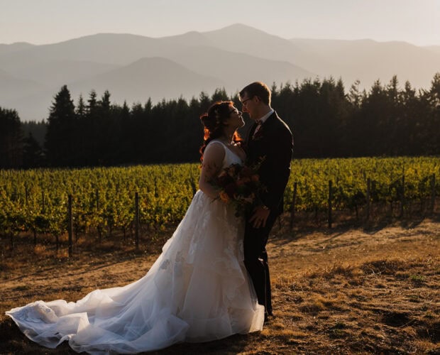 The bride and groom are embracing at Gorge Crest Vineyards with a sunset sky, mountains, and trees in the background.