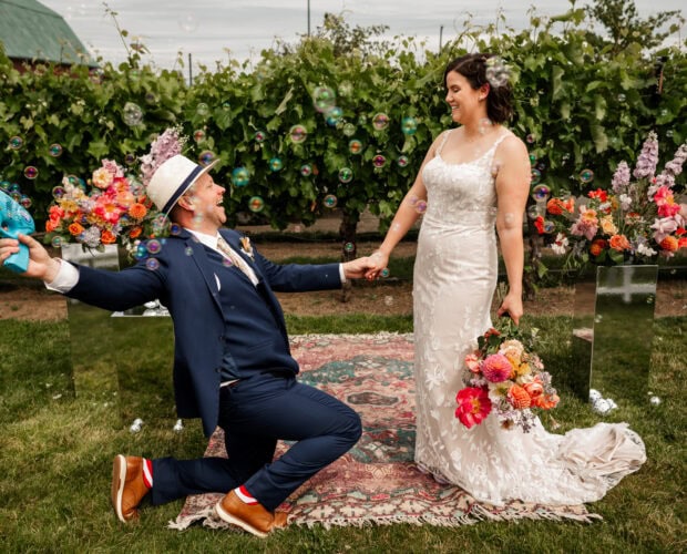 At Rossi Farms, the bride and groom pose in a vineyard with vibrant flowers around them. The groom kneels playfully with a bubble gun, adding a whimsical touch to their wedding photos.