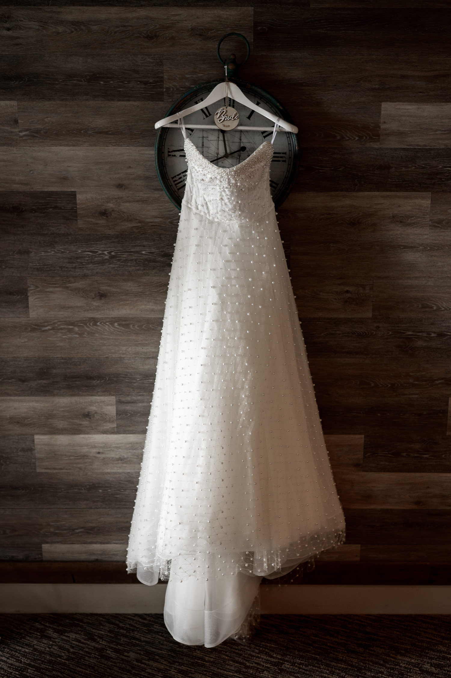 A white wedding dress with beaded details hangs on a clock-themed hanger against a wooden wall, preparing for a wedding at The Yachats Inn.