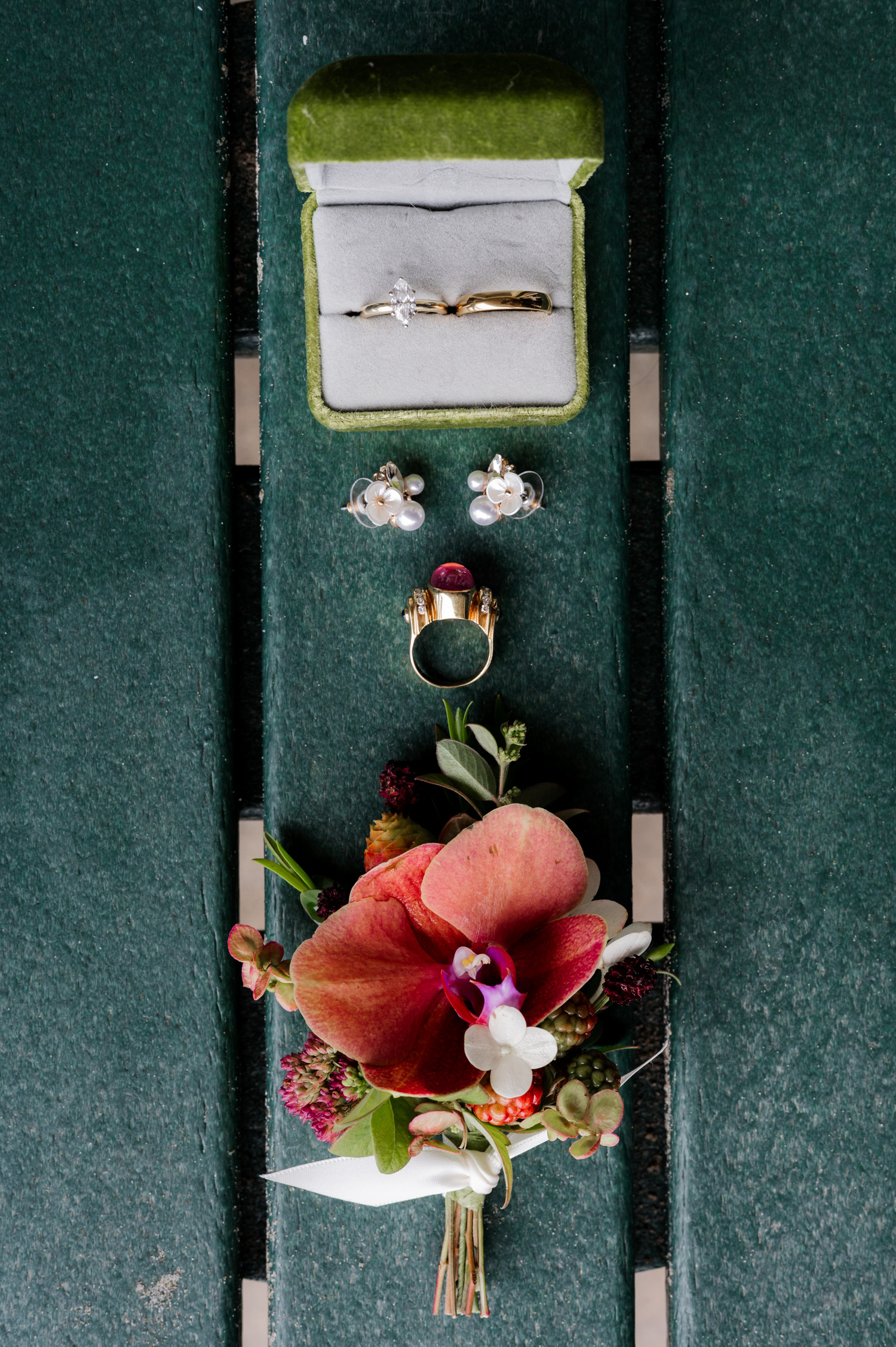 Wedding rings in a green box, floral-patterned earrings, and a bouquet with red and white flowers placed on a bench.