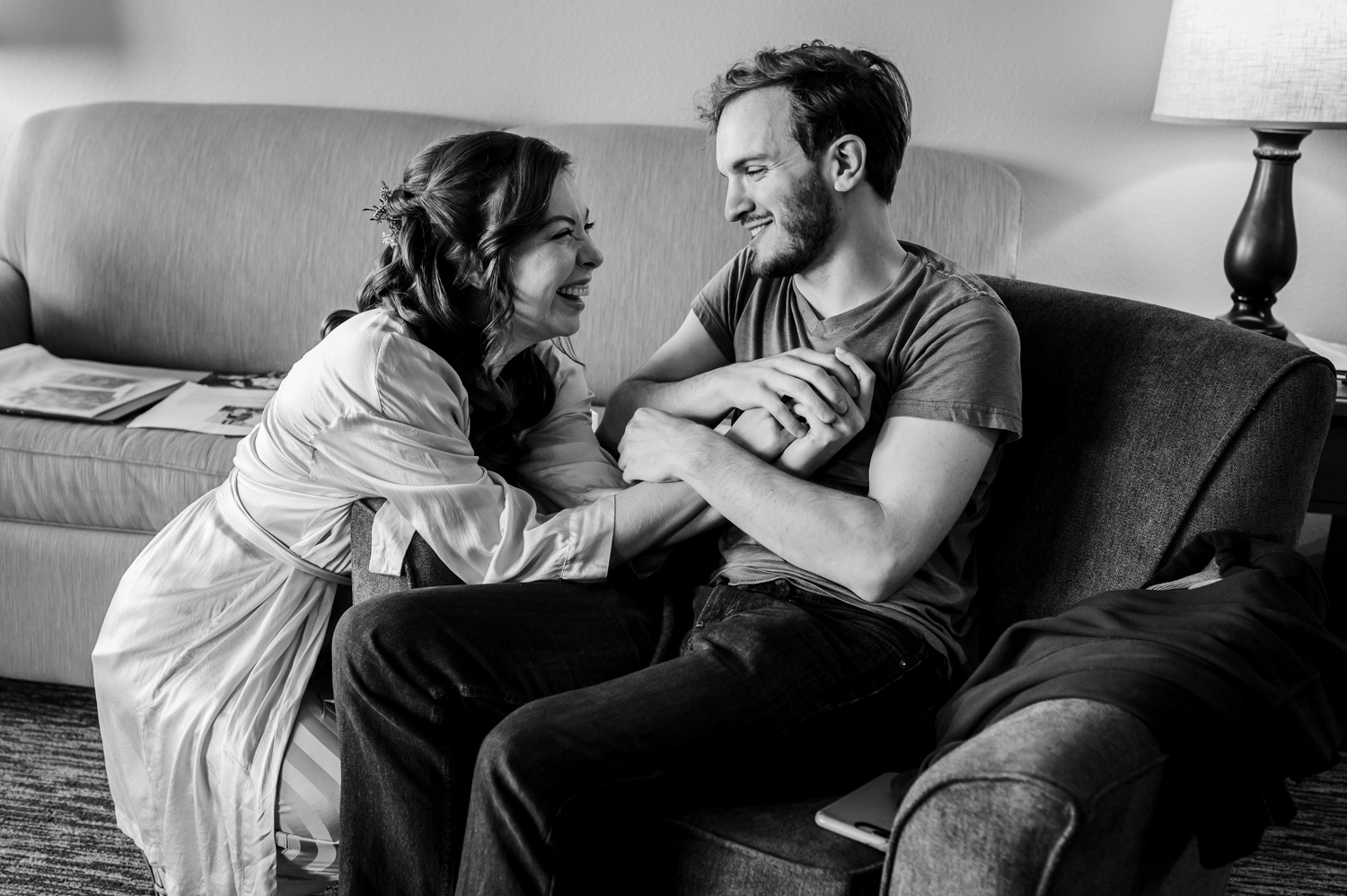 A woman in a robe smiles and holds hands with a man sitting in a chair, sharing a joyful moment as they prepare for their wedding at The Yachats Inn.