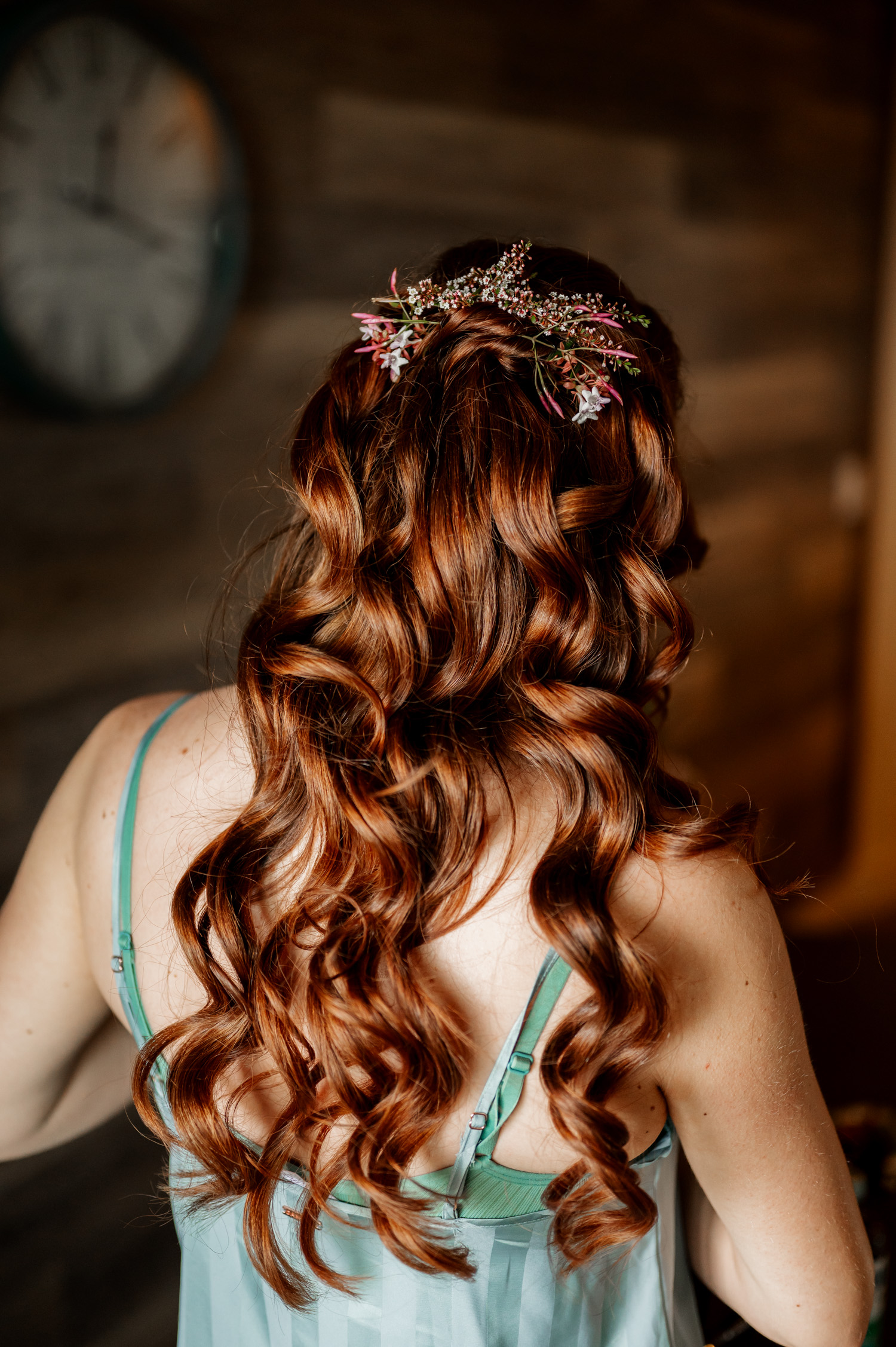 A woman with long, curly red hair and a floral hairpiece is in a teal tank top, preparing for a wedding. She is facing away while getting ready at the Overleaf Lodge and Spa.