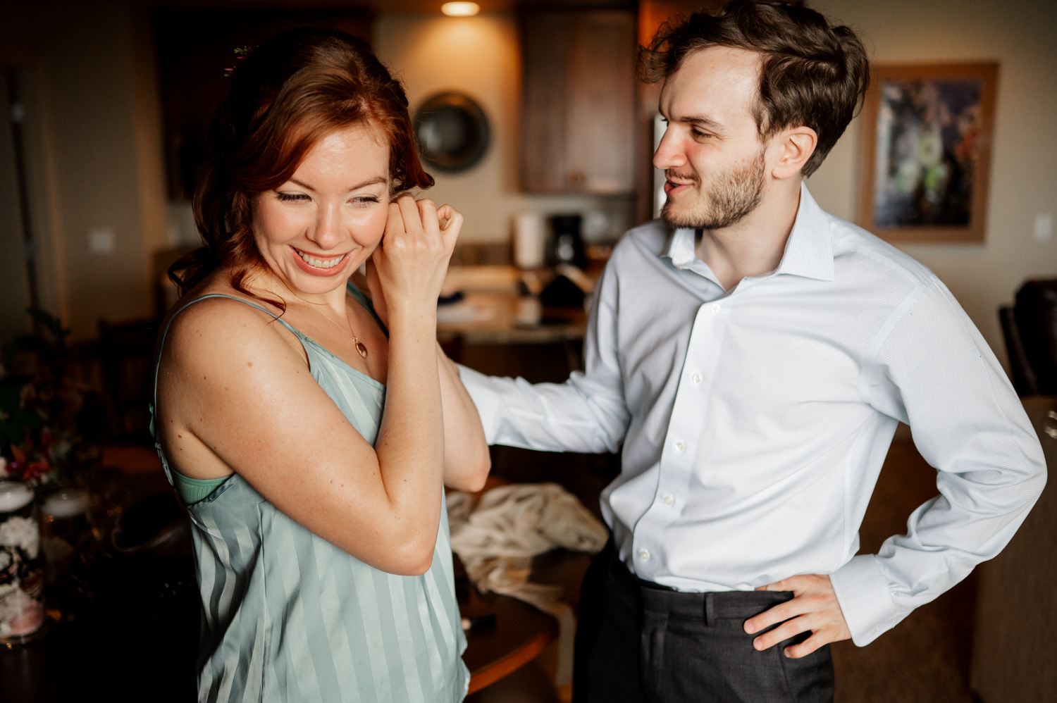 A woman smiling as she adjusts her earring stands next to a man in a white shirt indoors, both preparing for a wedding celebration.