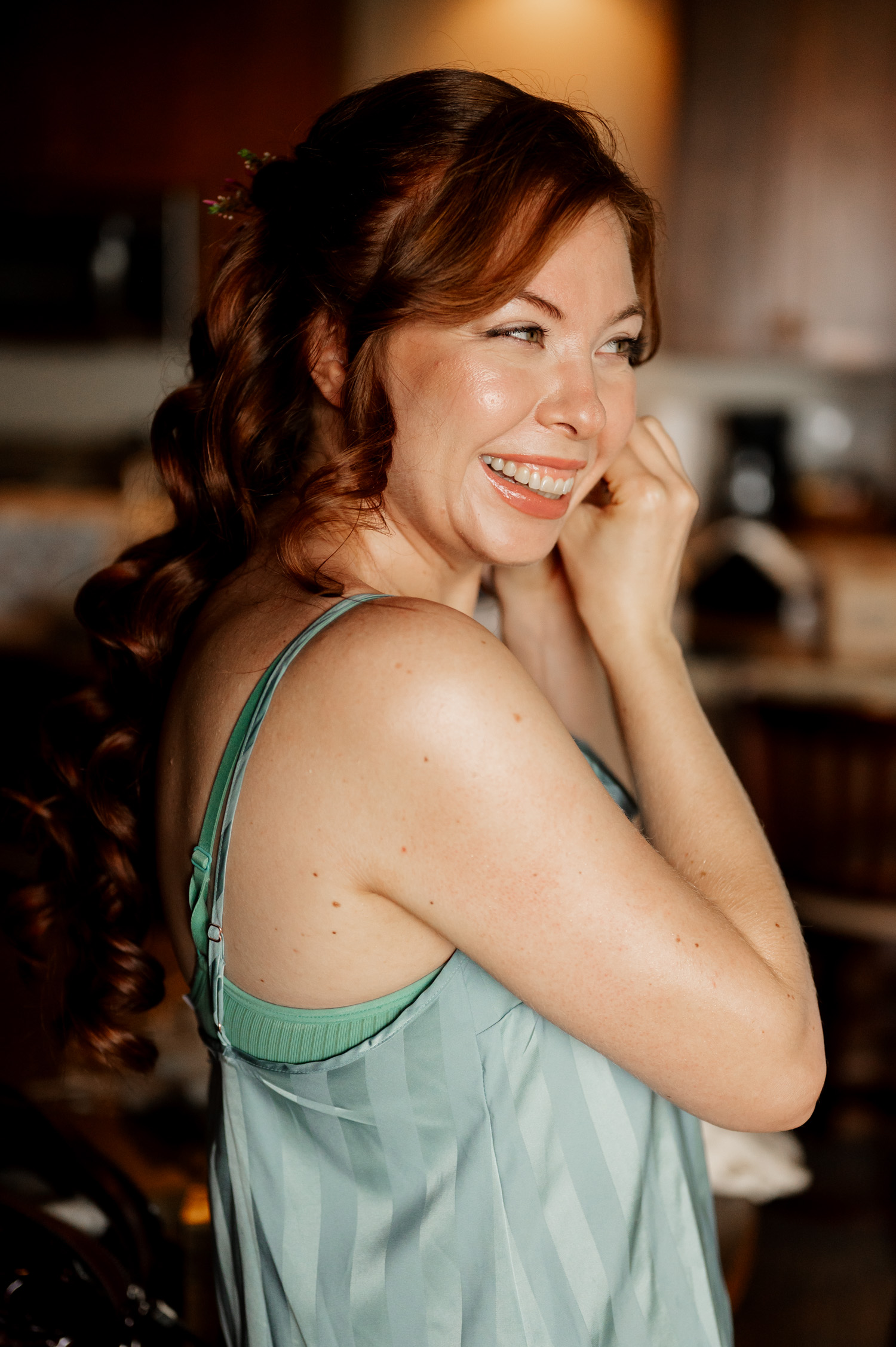 In the cozy Overleaf Lodge and Spa, a smiling woman with long red hair adjusts her earring while wearing a striped green top. She is preparing for a wedding at The Yachats Inn.