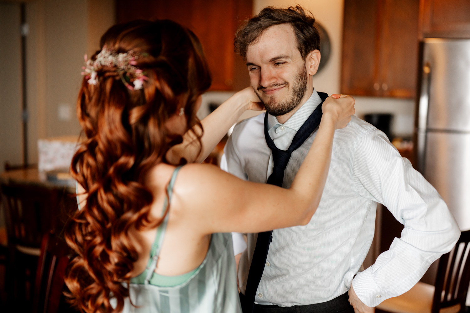 A woman adjusts a mans tie in the kitchen, both smiling warmly. They are preparing for their wedding day at The Yachats Inn, staying at the Overleaf Lodge and Spa.