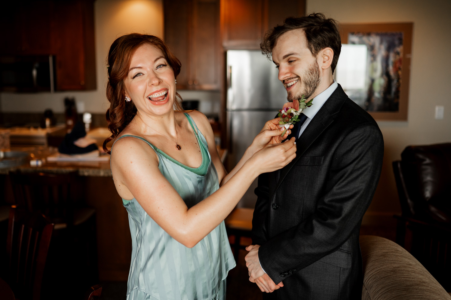 A woman in a green dress pins a boutonniere on a smiling man in a dark suit in a cozy kitchen setting. Theyre preparing for their wedding at The Yachats Inn, staying at Overleaf Lodge and Spa.