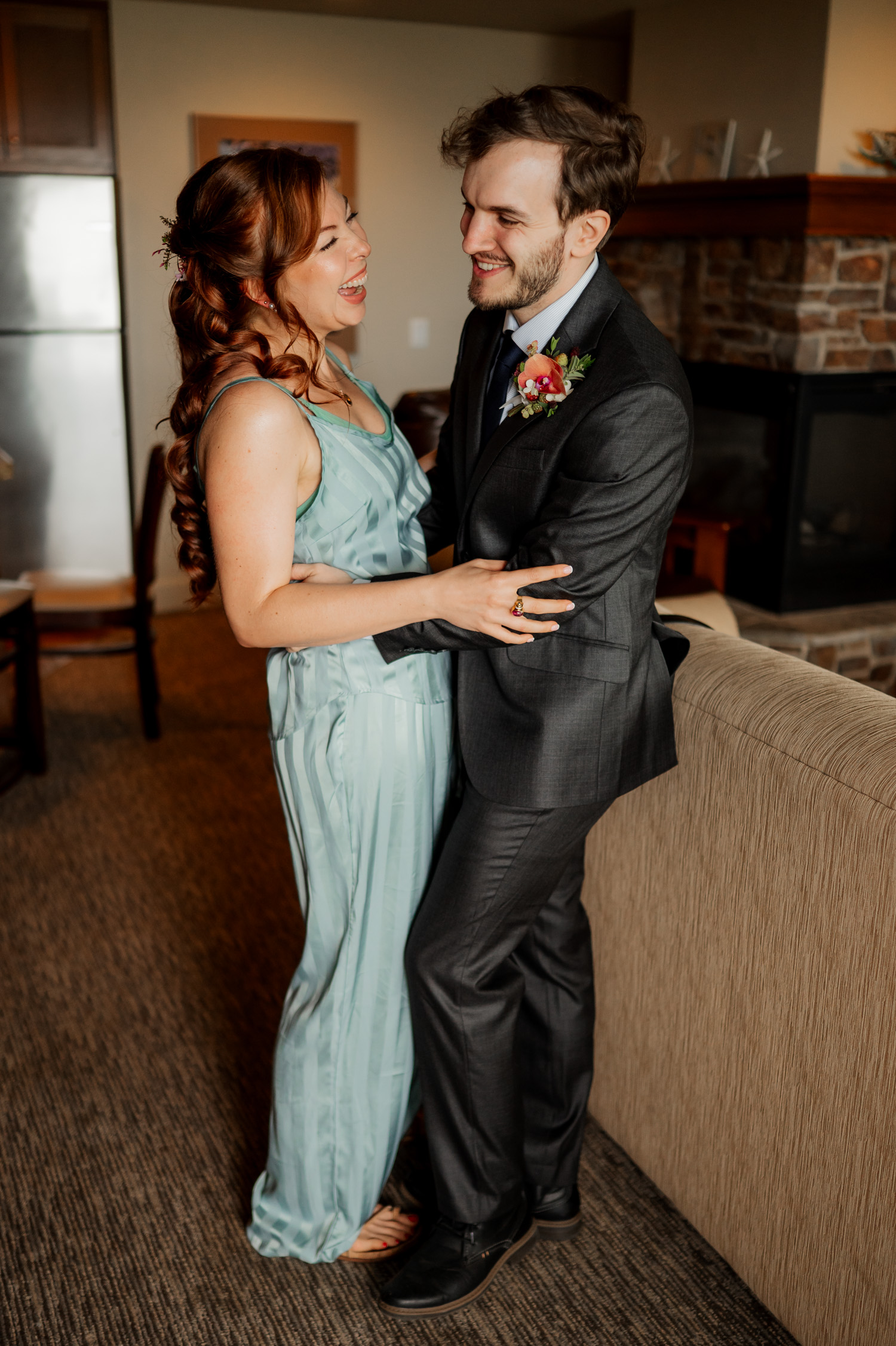 A couple embracing, smiling, and laughing in a cozy living room. The woman is wearing a turquoise dress and the man is in a suit. They are preparing for their wedding at The Yachats Inn while at the Overleaf Lodge and Spa.