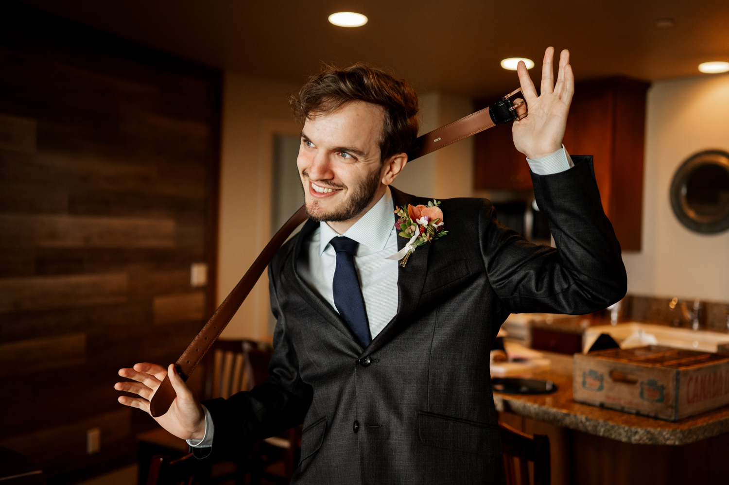 A man in a suit smiles while holding a belt over his shoulder, featuring a boutonniere on his lapel. Hes at Overleaf Lodge and Spa, getting ready for a wedding at The Yachats Inn.