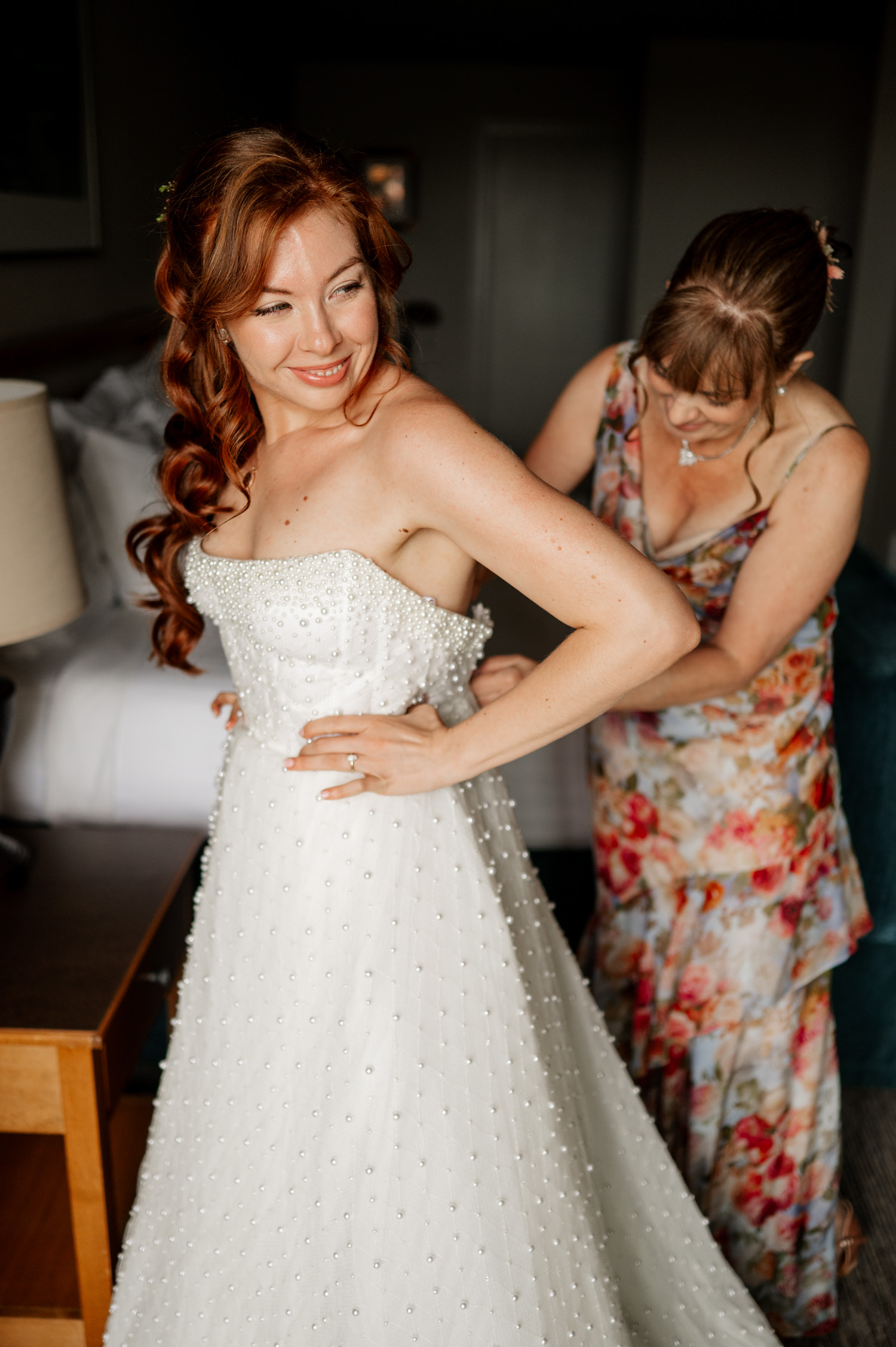 A bride smiles brightly in a pearl-studded gown while a woman in a floral dress assists her with adjustments. They are preparing for a wedding at Overleaf Lodge and Spa, set to take place at The Yachats Inn.