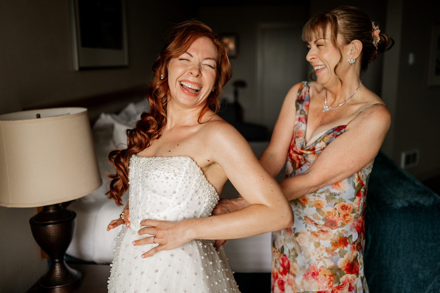 A bride, smiling broadly, stands in a warmly lit room at Overleaf Lodge and Spa as a woman in a floral dress assists her with adjusting her gown.