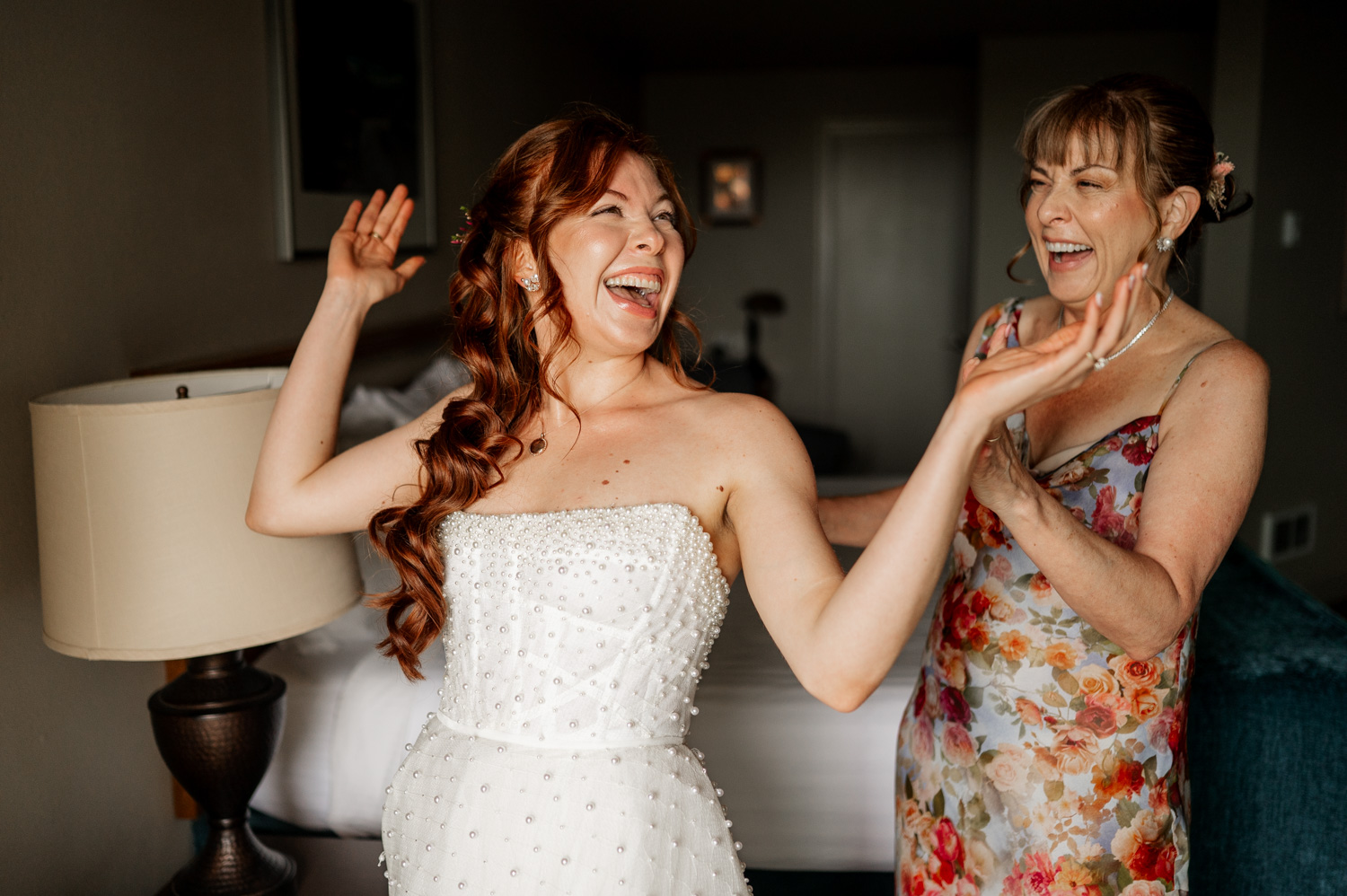 A joyful bride in a beaded dress laughs with a woman wearing floral attire in a warmly lit bedroom as they prepare for the wedding at The Yachats Inn, with the setting being at Overleaf Lodge and Spa.