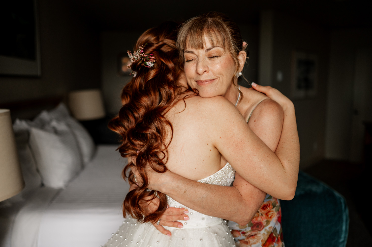 Two women share a warm embrace in a cozy bedroom at Overleaf Lodge and Spa. One wears a wedding dress, the other is in floral attire, both with eyes gently closed, cherishing the moment before leaving for The Yachats Inn celebration.