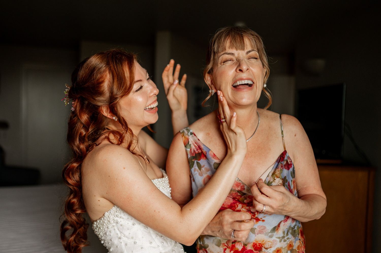 A bride and a woman in a floral dress share a joyful laugh while preparing for a wedding at the Overleaf Lodge and Spa.