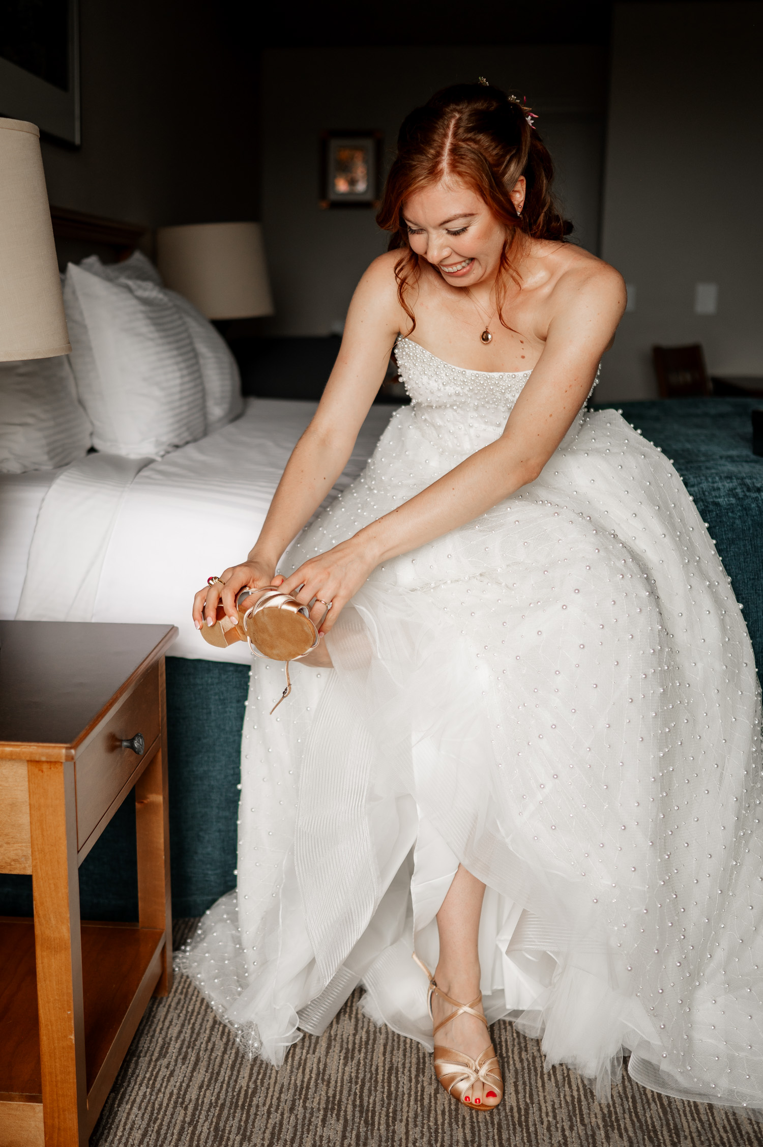 Bride in a white gown sitting on a bed, smiling as she puts on gold heels, preparing for her wedding.