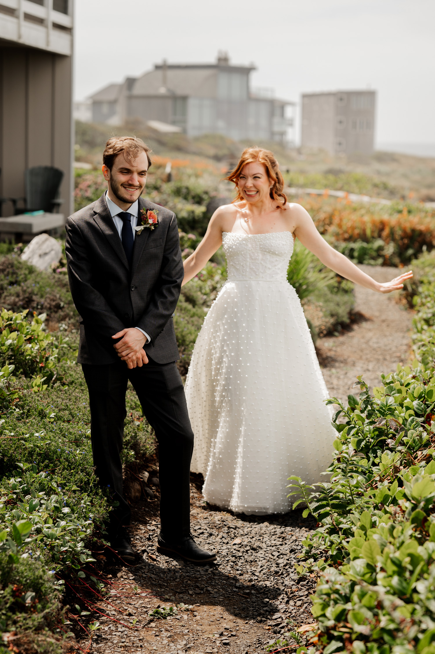 A bride in a white dress playfully surprises the groom, who is wearing a dark suit, on a charming garden path. Both are smiling joyfully, capturing a happy moment before their wedding celebration.