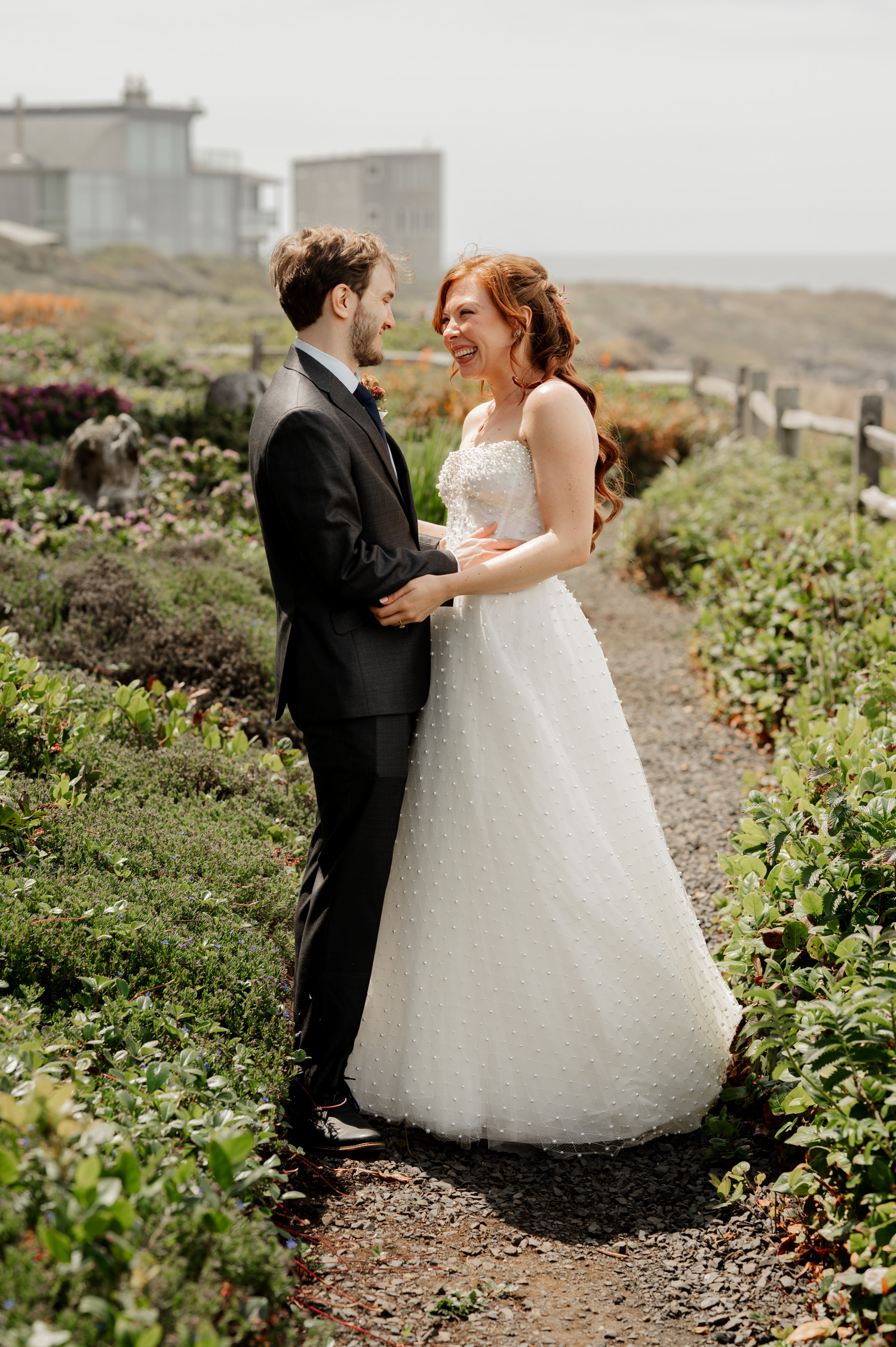 Bride and groom smiling at each other on a garden path with the ocean and houses in the background.