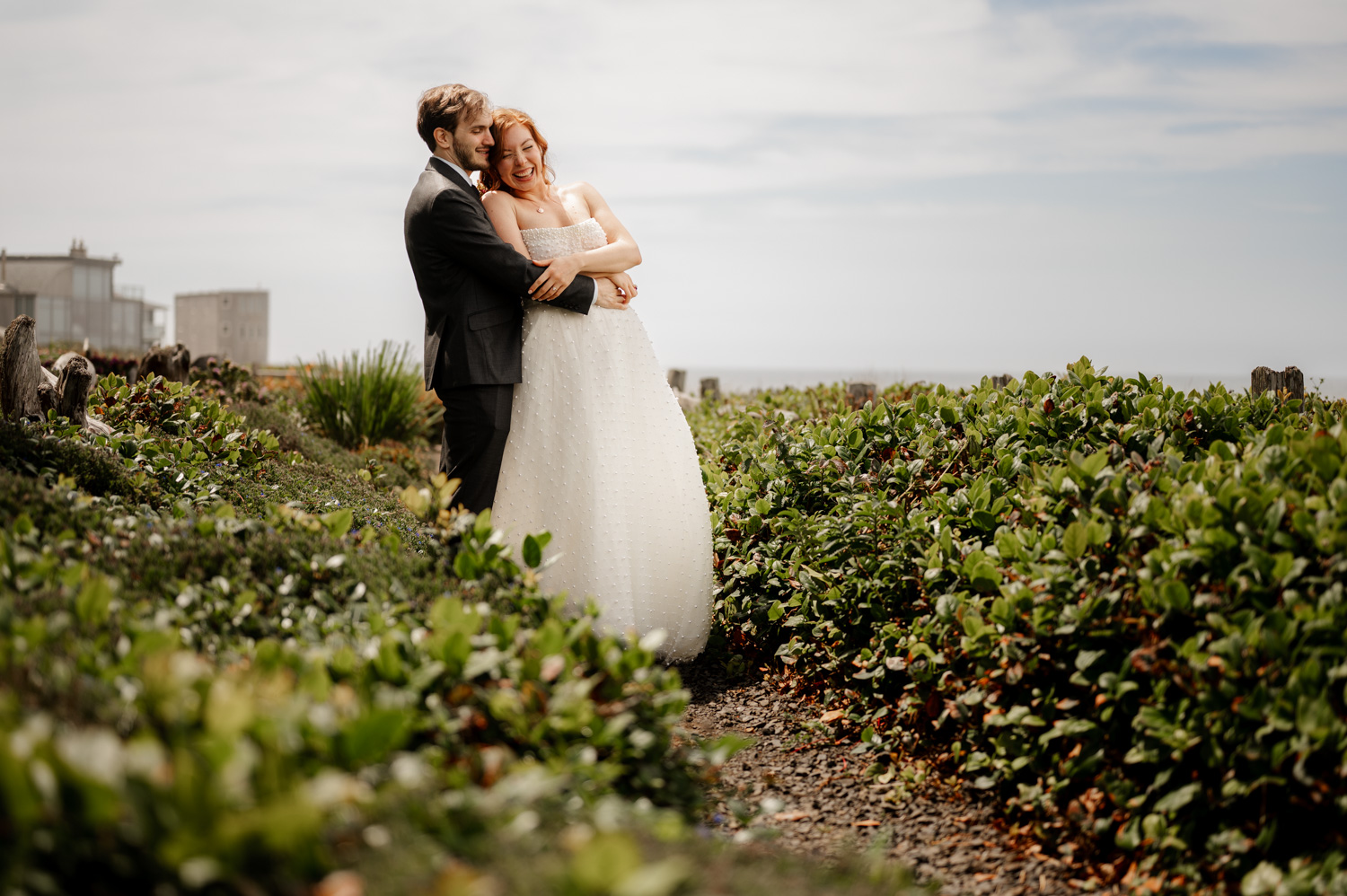 A bride and groom embrace in a lush garden, surrounded by greenery with a building in the background.