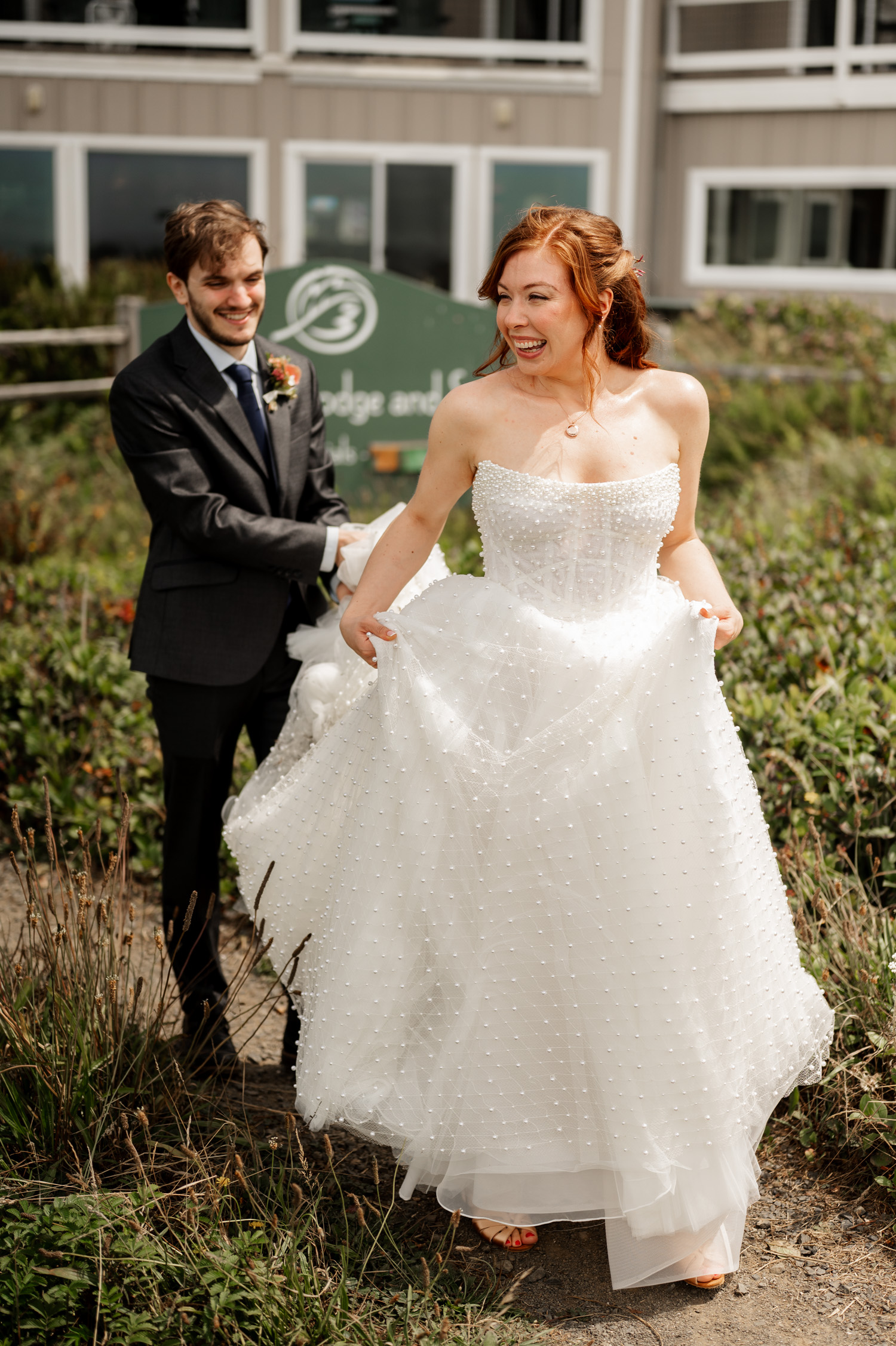 A bride holds her dress with a smile, while the groom stands behind her in his suit. They are surrounded by lush greenery near a charming building, capturing their joyful wedding portraits at the Overleaf Lodge and Spa.