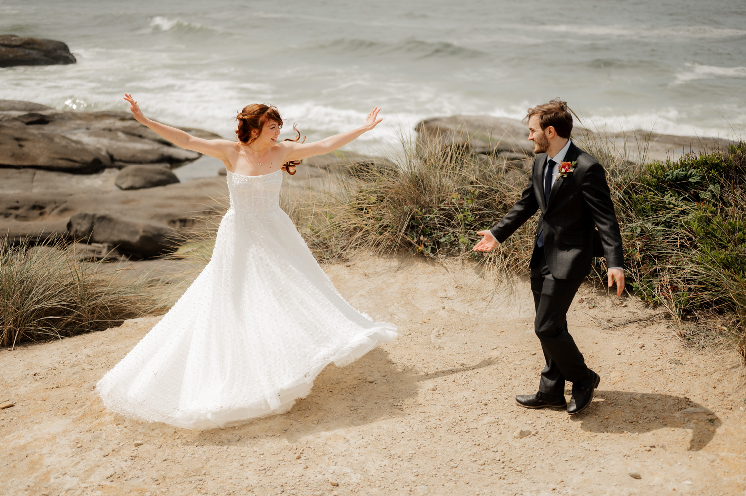Bride and groom joyfully dancing on a sandy beach with rocks and ocean waves in the background.
