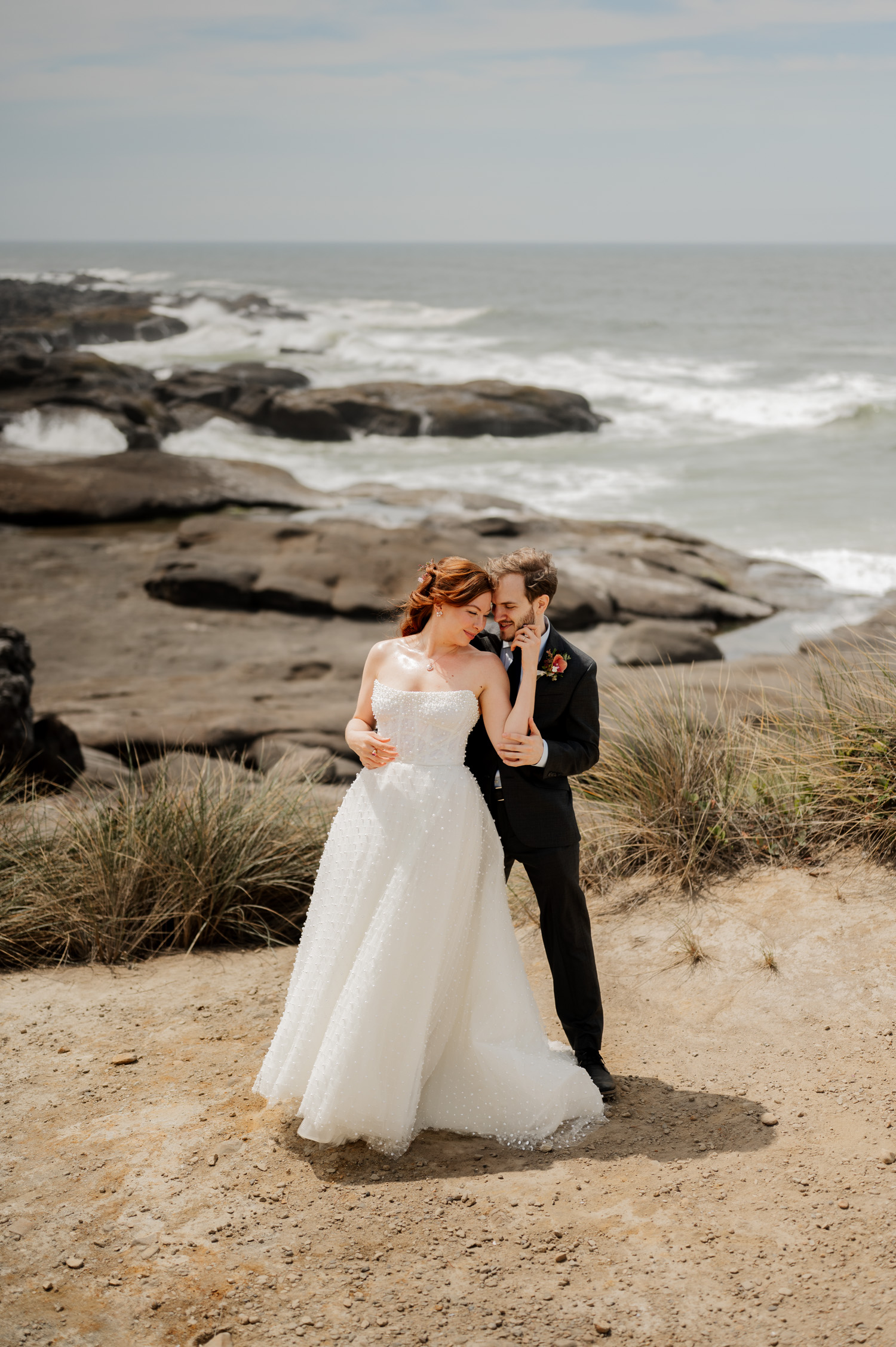 The bride and groom embrace on a rocky beach with waves crashing behind them. Their wedding portraits were taken at the Overleaf Lodge and Spa, capturing the serene beauty before their union at the Yachats Inn.