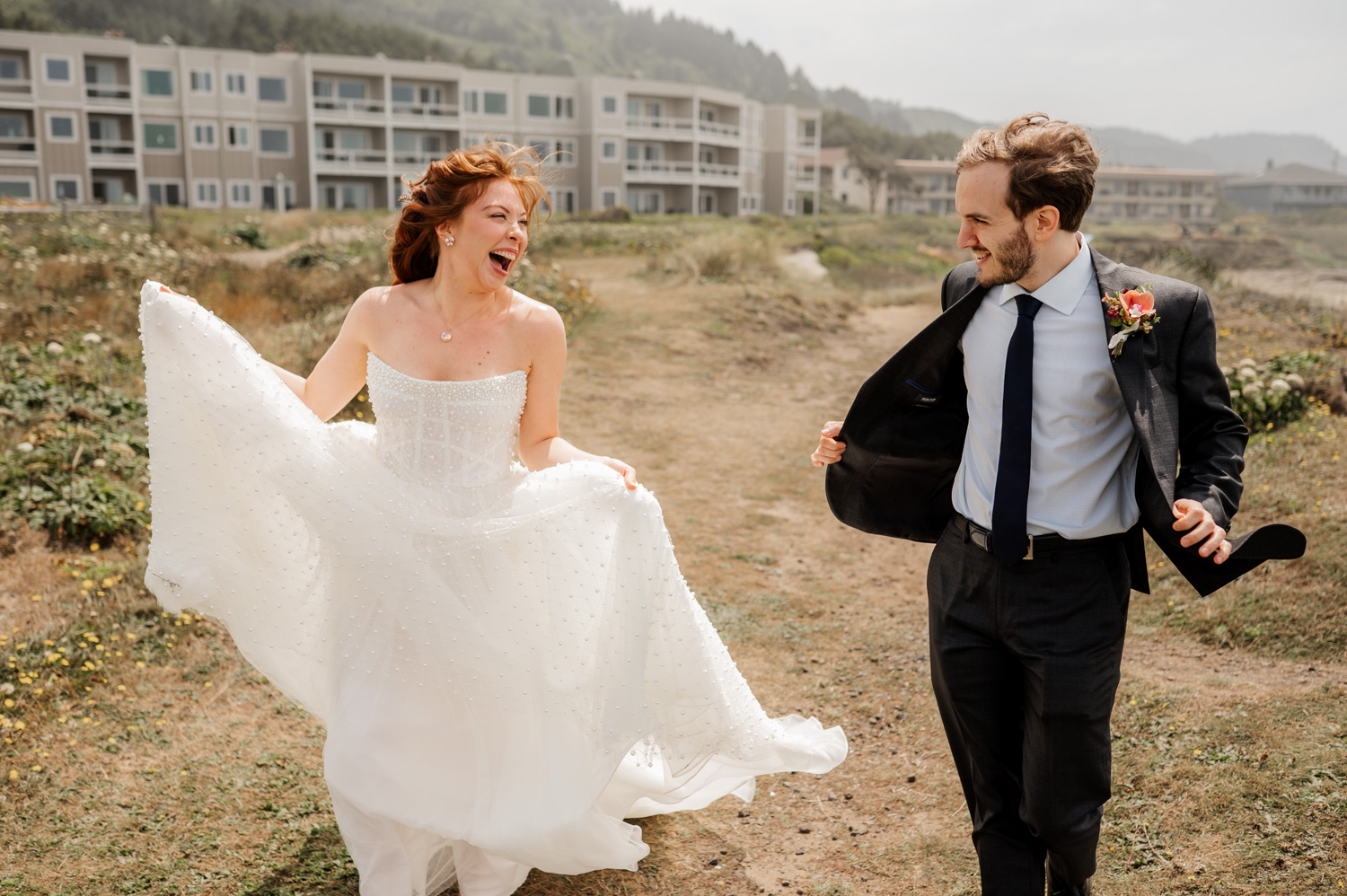 A joyful couple running outdoors on their wedding day, surrounded by a grassy landscape and buildings in the background.