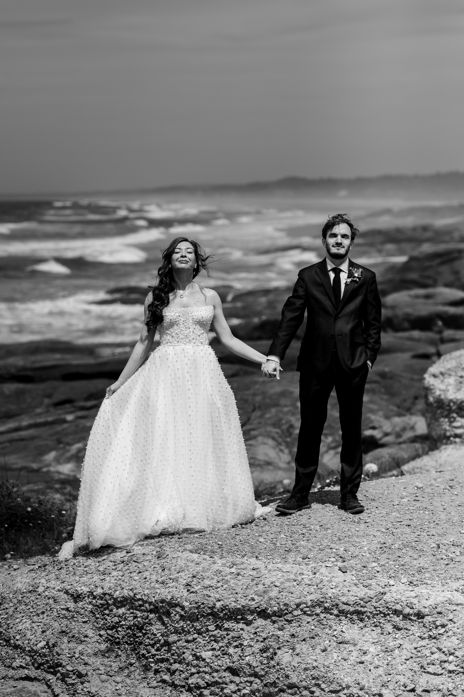 A bride and groom stand on a rocky shore, holding hands and smiling, with waves crashing in the background. They are at the Overleaf Lodge and Spa before their wedding at the Yachats Inn.