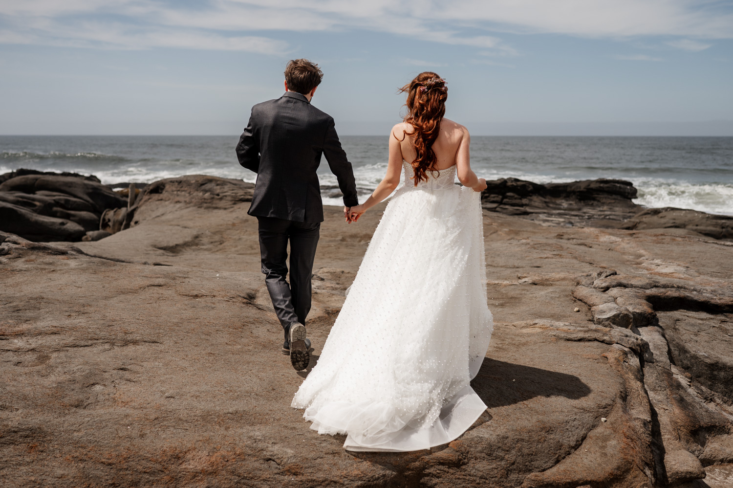 Bride and groom holding hands while walking on the rocky shore with the ocean in the background.