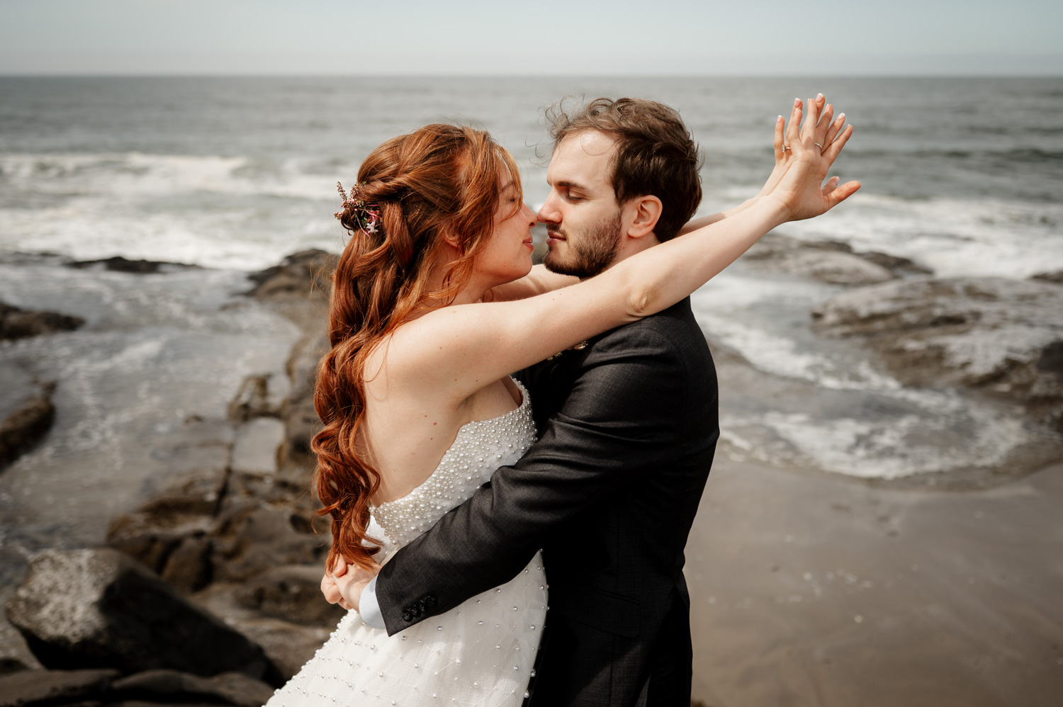 A couple embraces on a rocky beach near the Overleaf Lodge and Spa with waves crashing behind. The woman has her arms raised joyfully as they celebrate their upcoming wedding at the Yachats Inn.