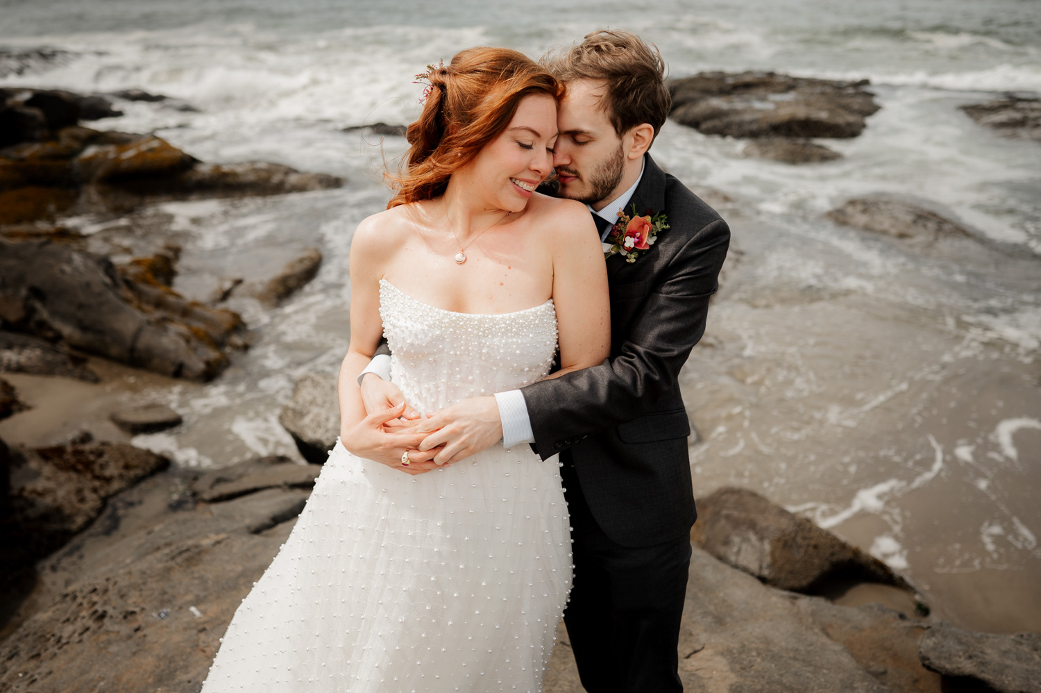 The bride and groom embrace on the rocky shore with ocean waves crashing in the background.