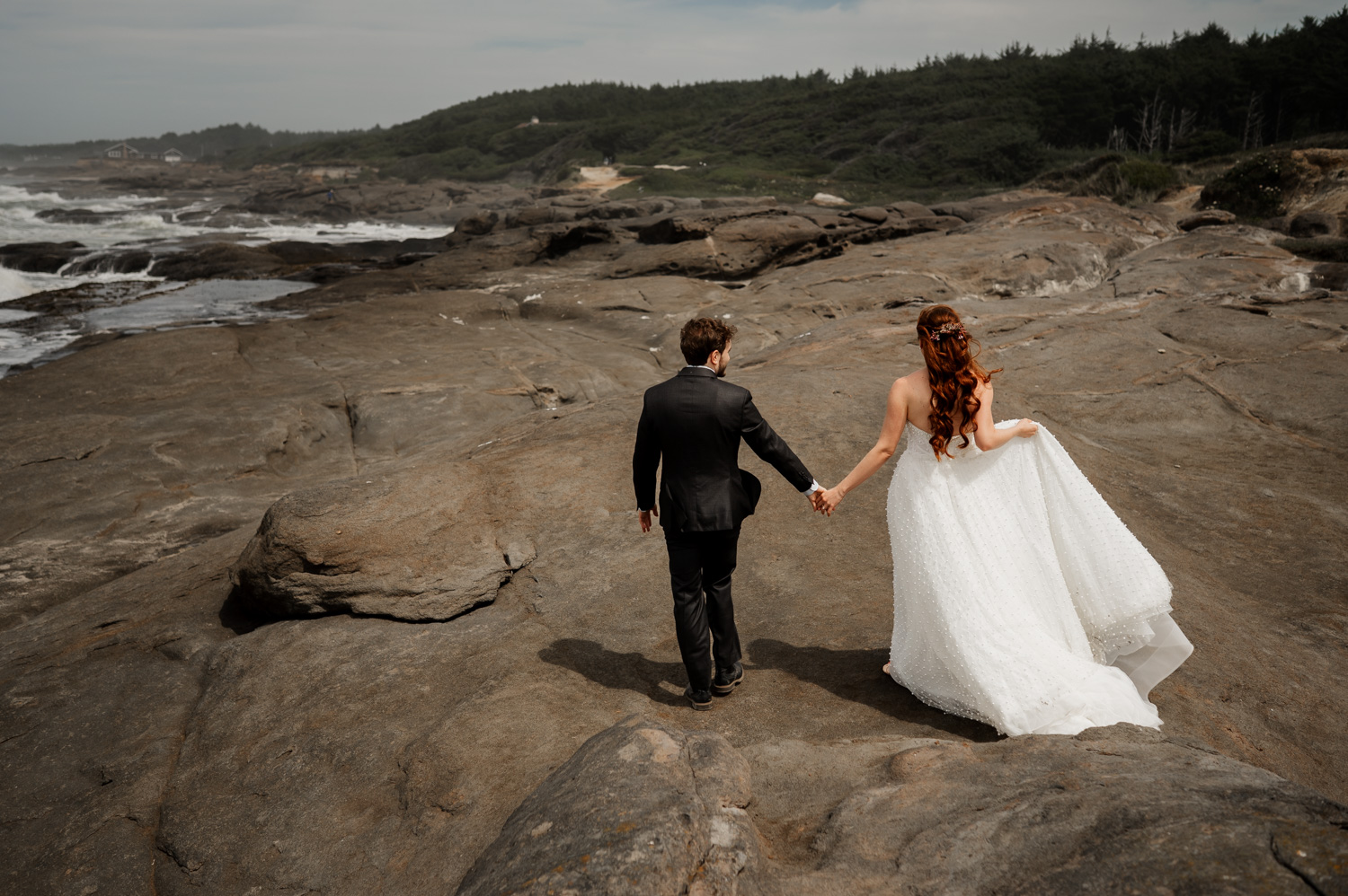 A bride and groom hold hands while walking on rocky terrain near a coastline, with trees in the background. They are capturing intimate wedding portraits at the Overleaf Lodge and Spa before their ceremony at the Yachats Inn.