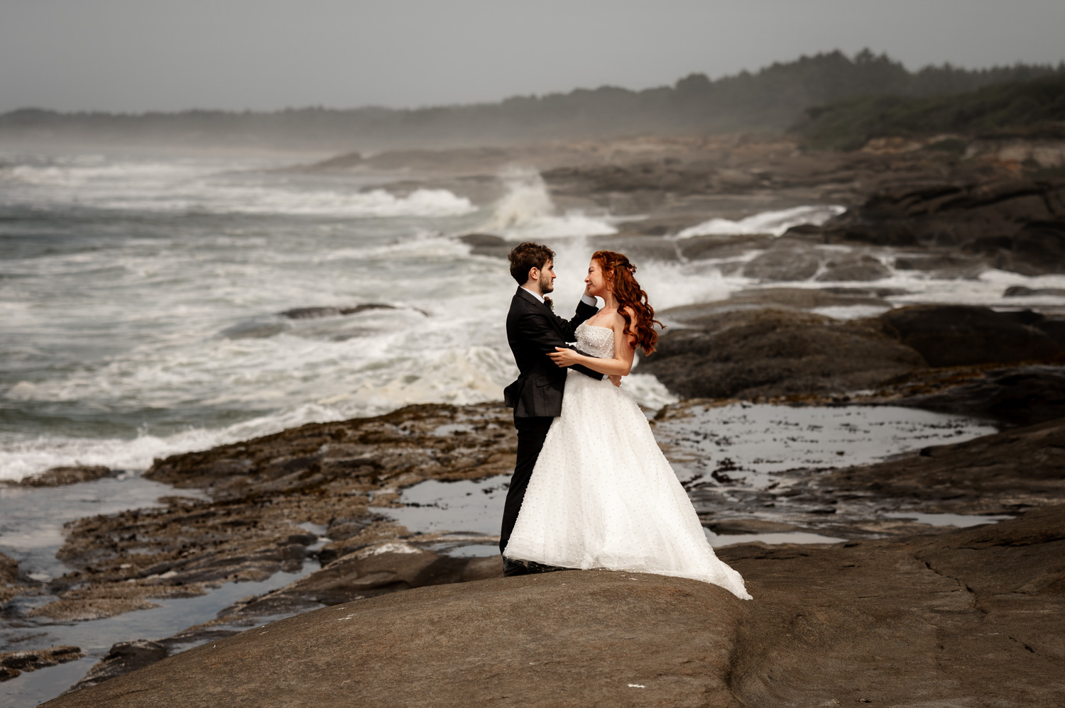 Bride and groom standing on a rocky shore with ocean waves in the background, taking wedding portraits at the Overleaf Lodge and Spa before their wedding at the Yachats Inn.