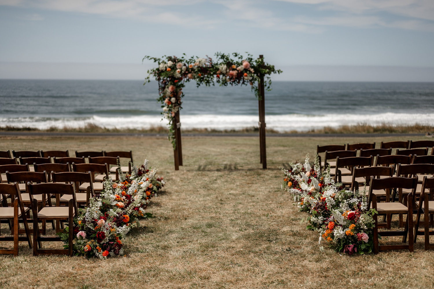An outdoor wedding setup by the sea at the Yachats Inn, featuring rows of chairs facing an archway adorned with flowers, with the ocean and a clear sky in the background.