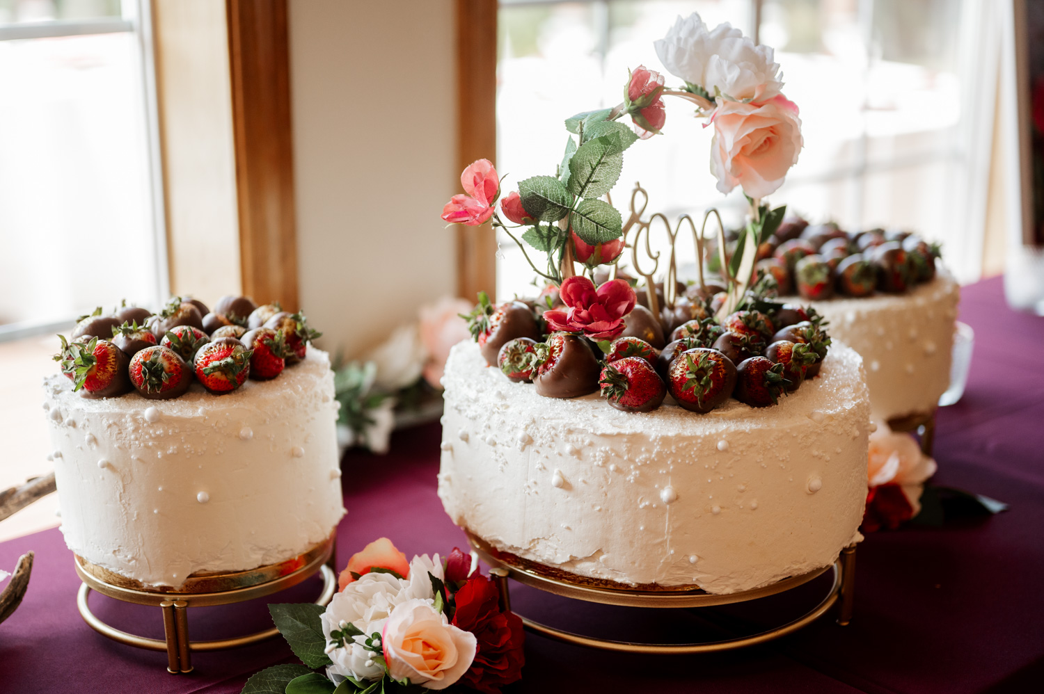 Three tiered cakes adorned with chocolate-covered strawberries and flowers are displayed on a burgundy cloth at the Yachats Inn.