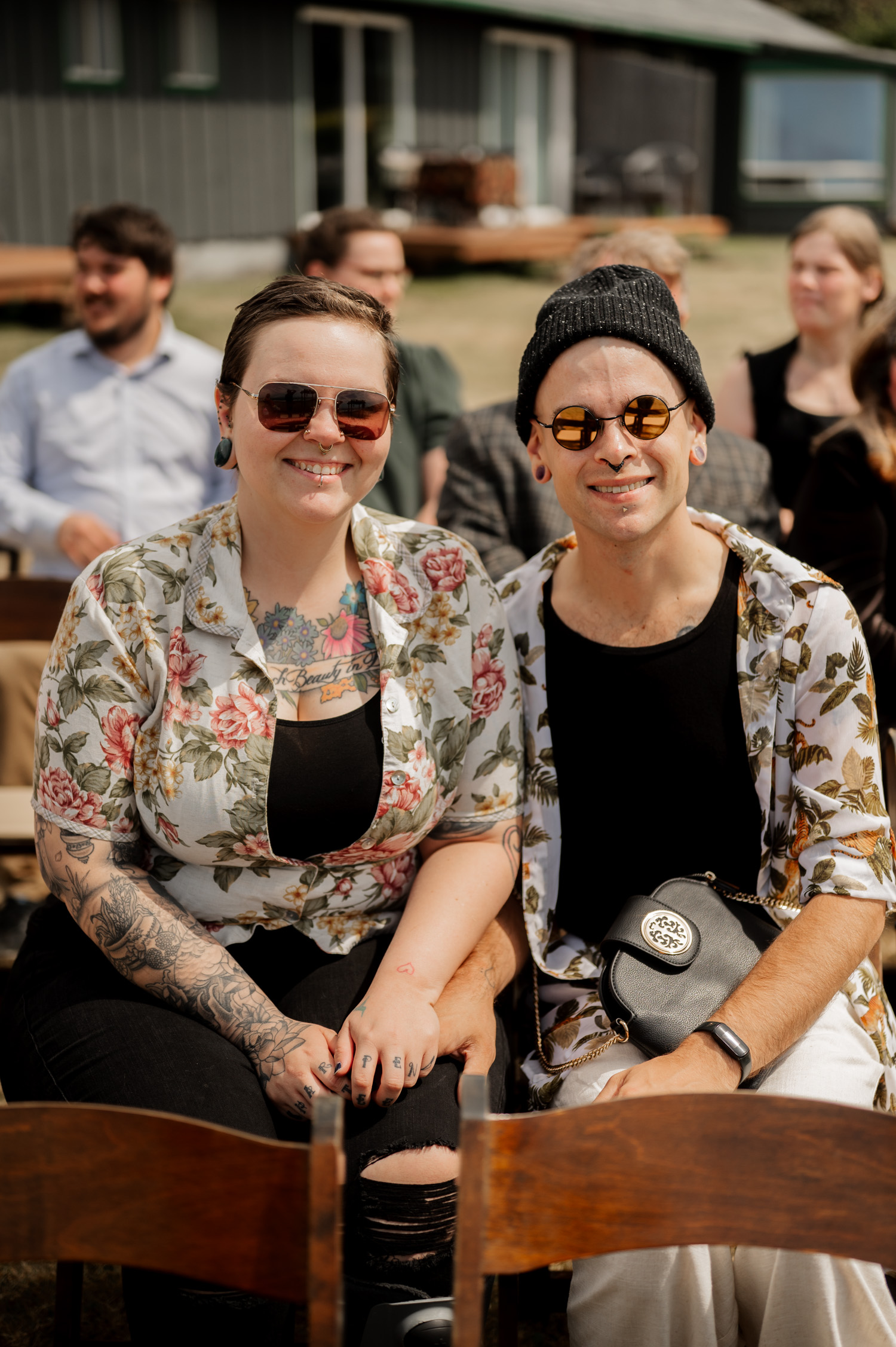 Two people in floral shirts and sunglasses smile while sitting outdoors at the Yachats Inn during a wedding. In the background, more guests are seated, contributing to a lively scene.
