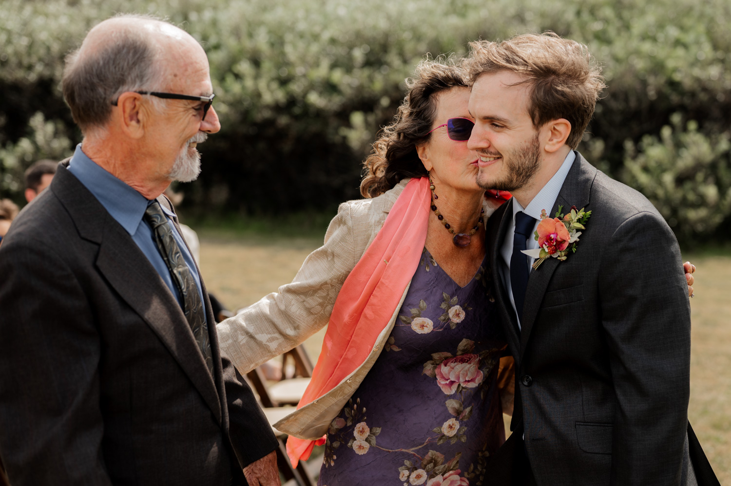An older couple embraces a younger man in a suit at an outdoor event, sharing a heartfelt moment among the wedding photos at the Yachats Inn.