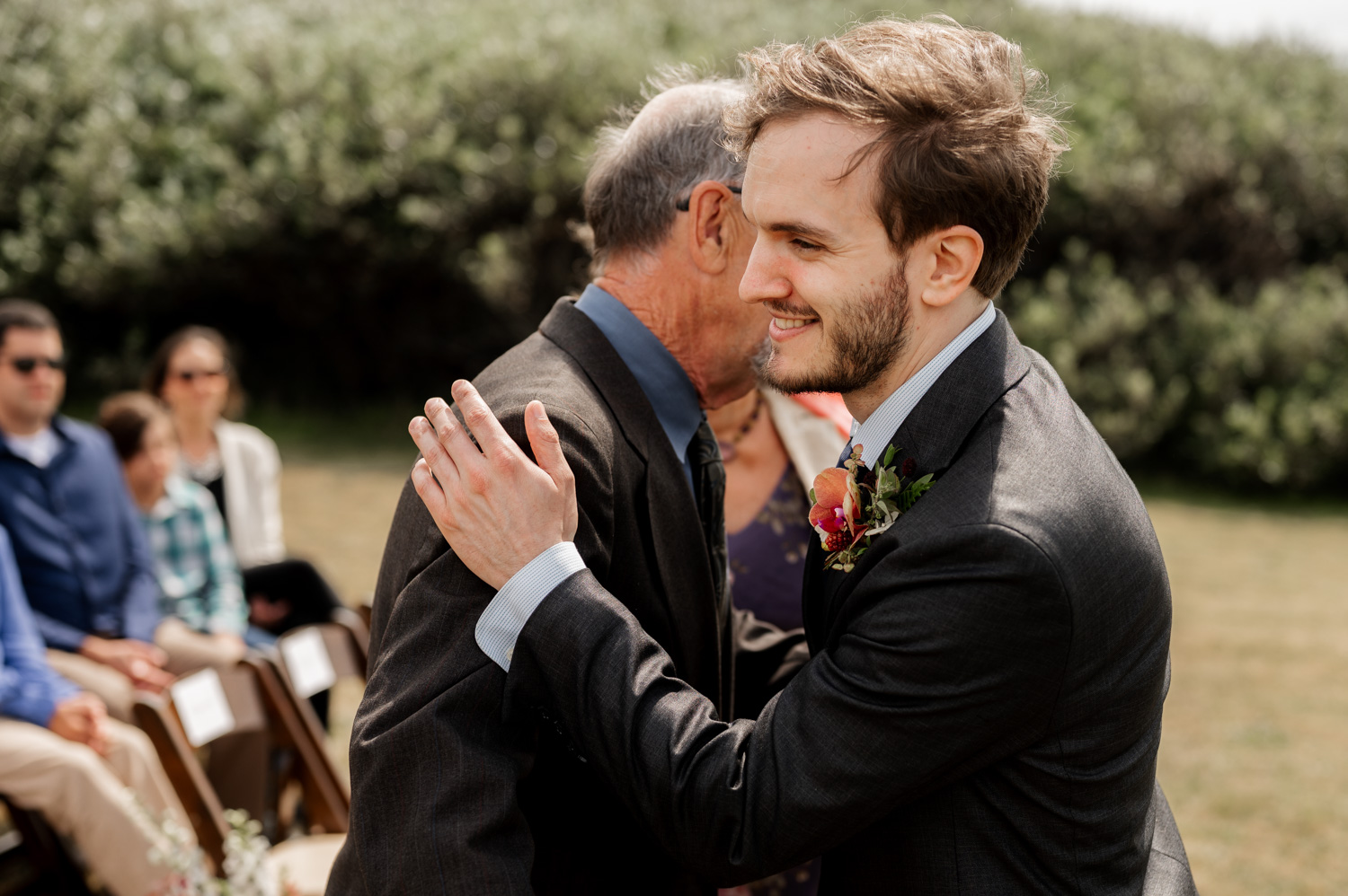 A man in a suit warmly embracing another man at an outdoor event, with seated guests visible in the background.