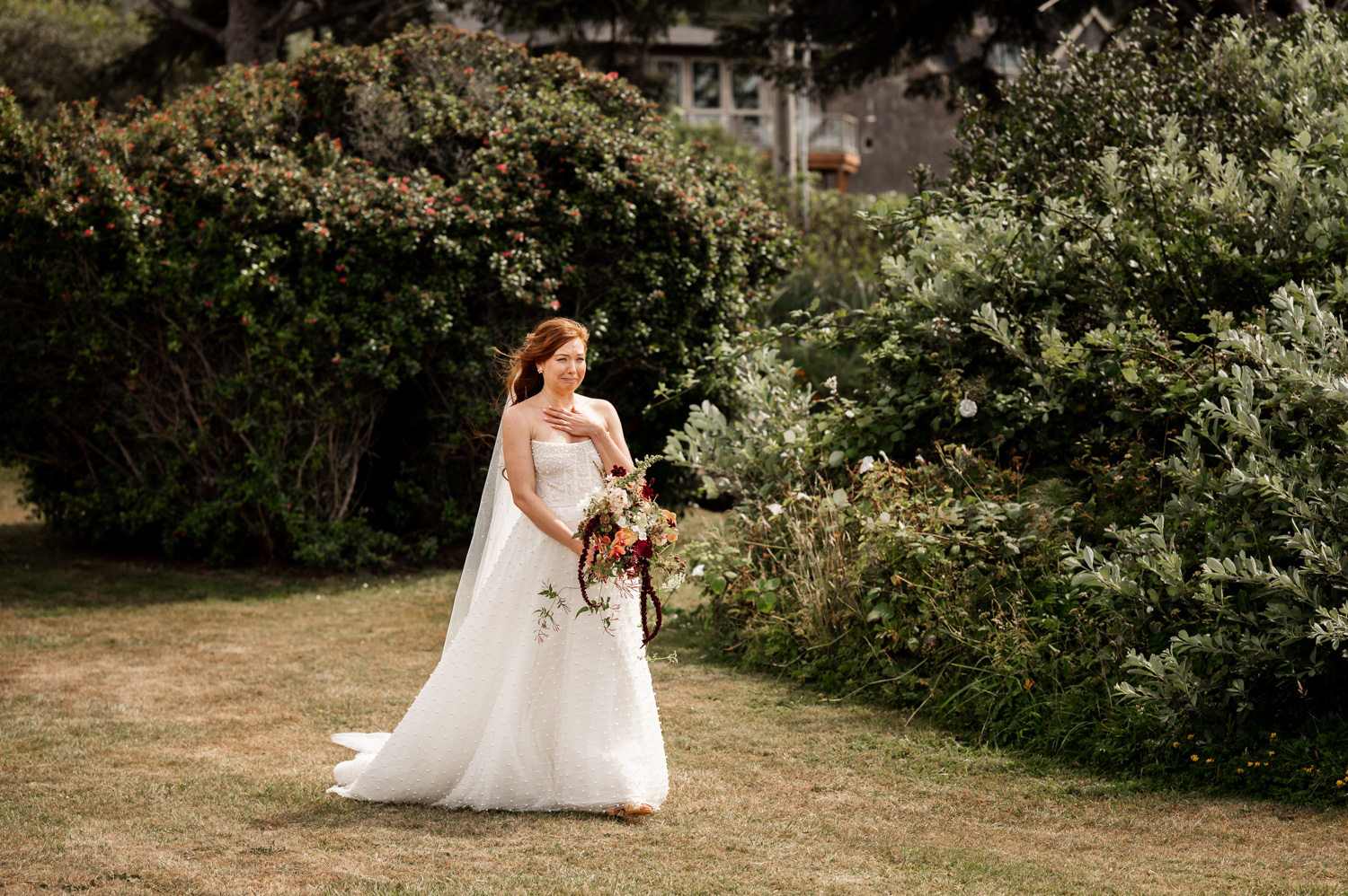 A bride in a white dress holding a bouquet walks gracefully across the grass, with lush greenery surrounding her at the Yachats Inn.