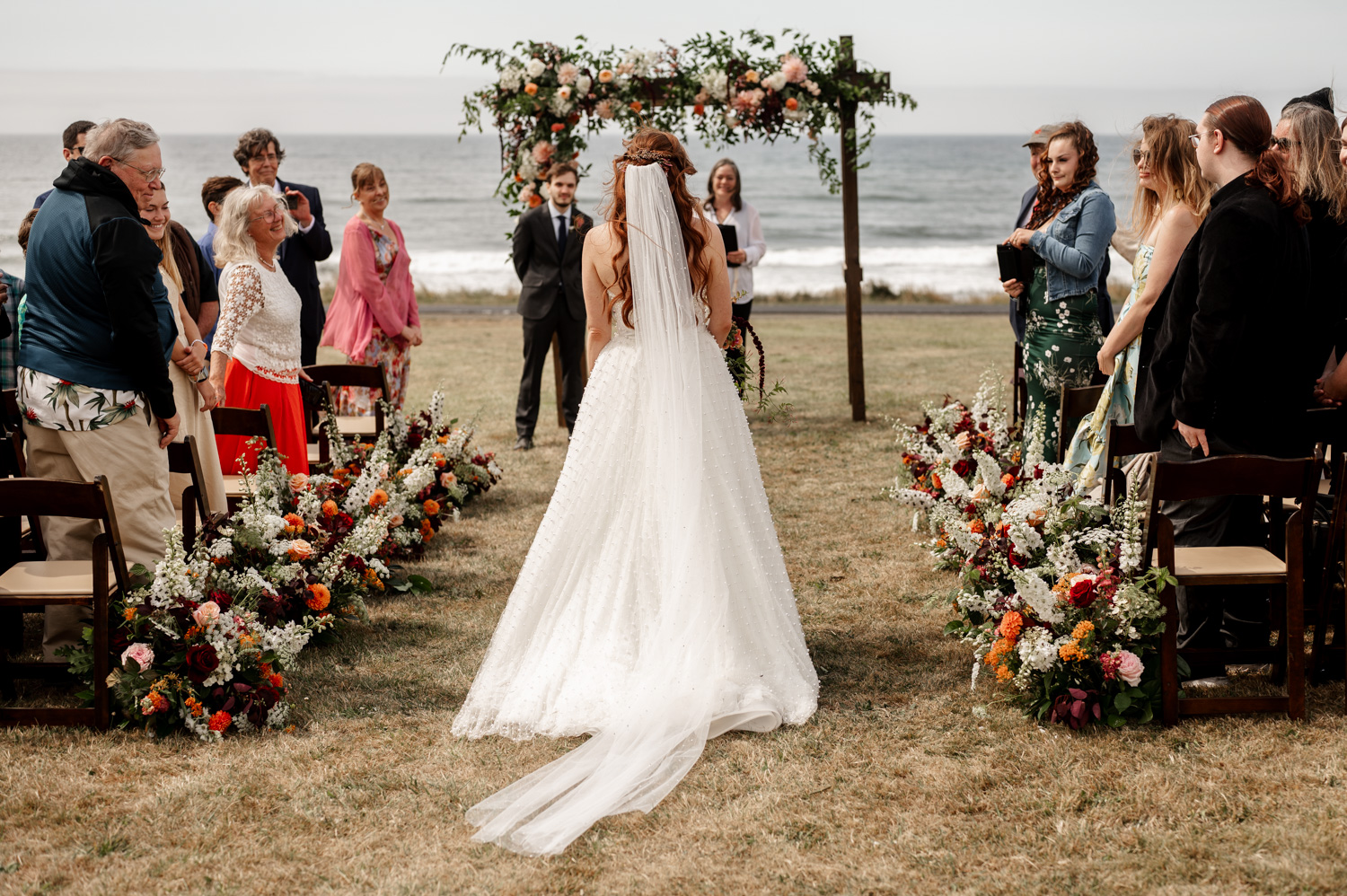 A bride walks down the aisle surrounded by elegant flower arrangements, with guests seated on either side and a stunning ocean backdrop at the Yachats Inn.