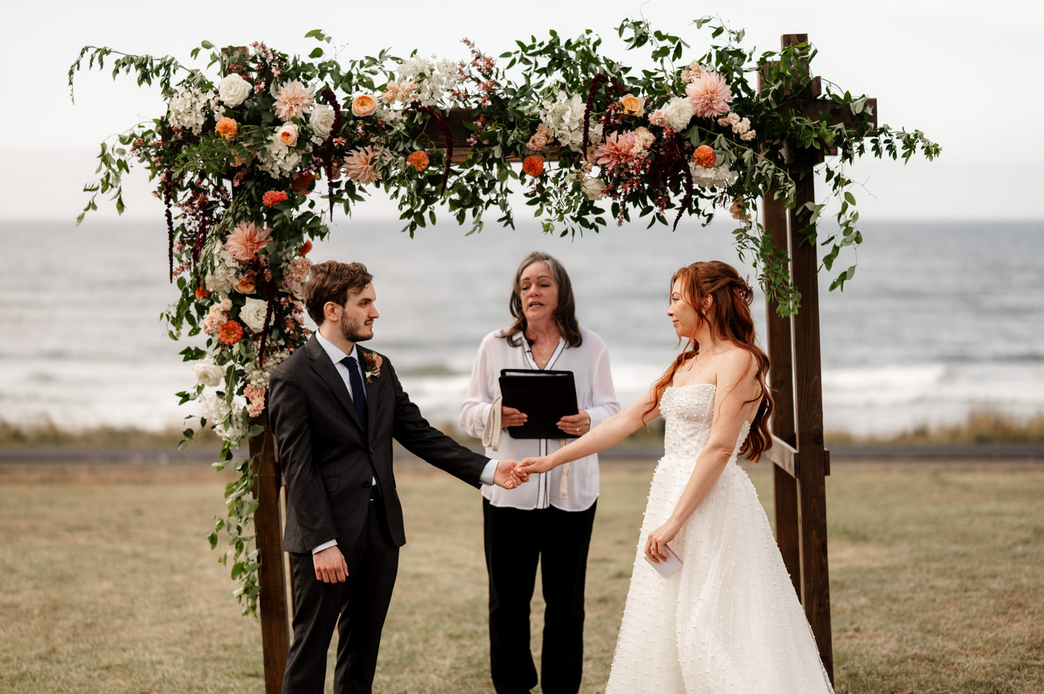 A couple holds hands during their wedding ceremony by the ocean, standing under a floral arch with an officiant at the Yachats Inn.