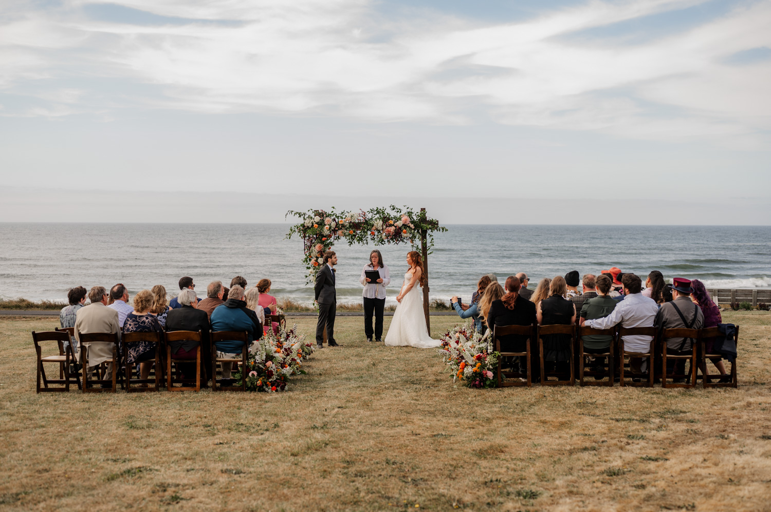 An outdoor beach wedding ceremony with a flower-covered arch, guests seated in rows on both sides, and the ocean visible in the background.