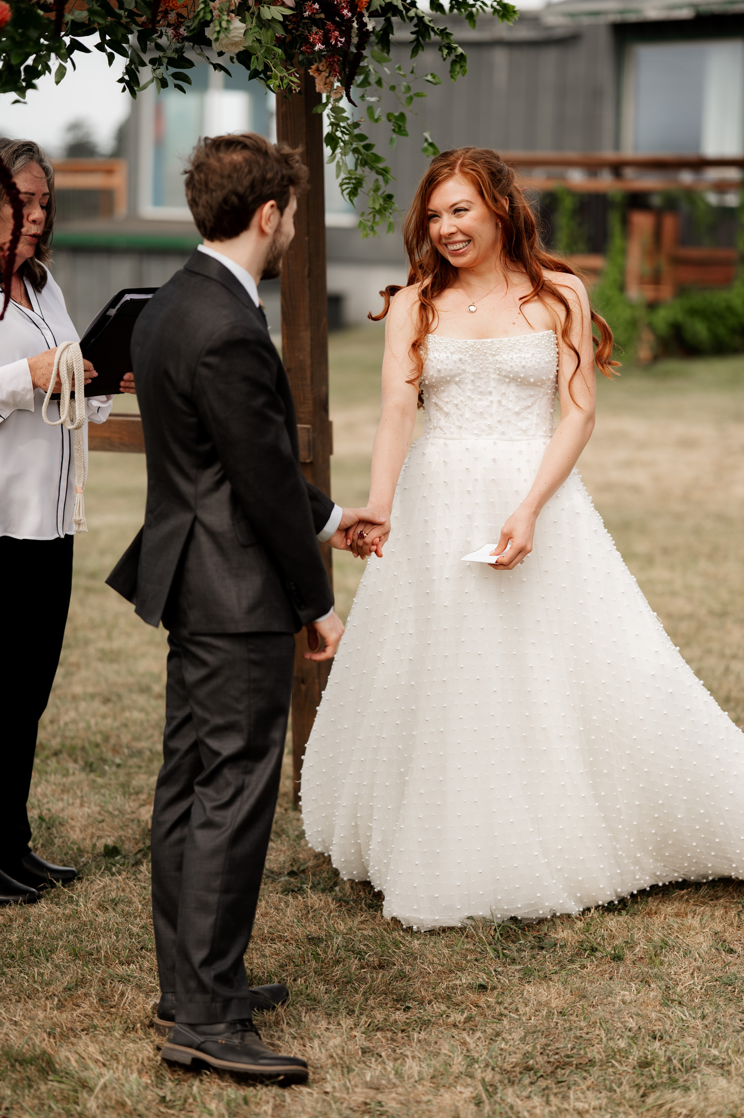 Bride and groom exchange smiles while holding hands during an outdoor wedding ceremony with a serene backdrop at the Yachats Inn.