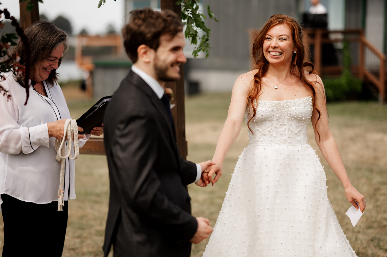 A bride in a white dress and groom in a dark suit hold hands, smiling joyfully during their outdoor wedding ceremony at the Yachats Inn.