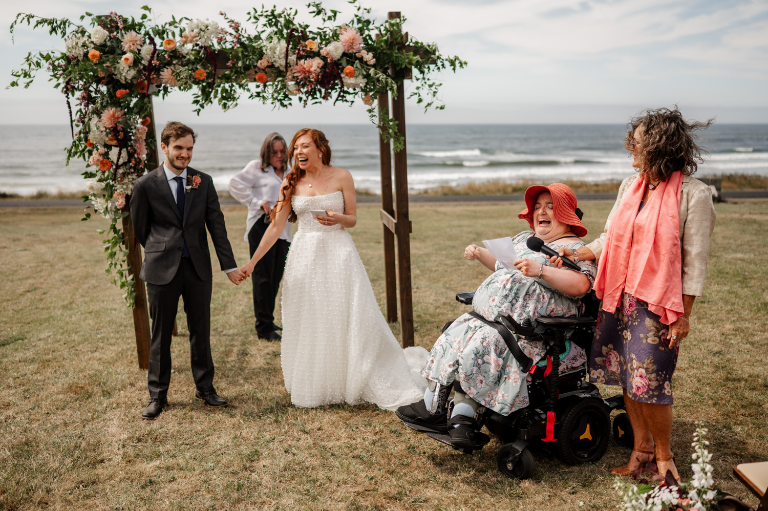 A smiling couple stands under a floral arch during a joyful wedding ceremony by the ocean, with a guest in a wheelchair wearing a red hat.
