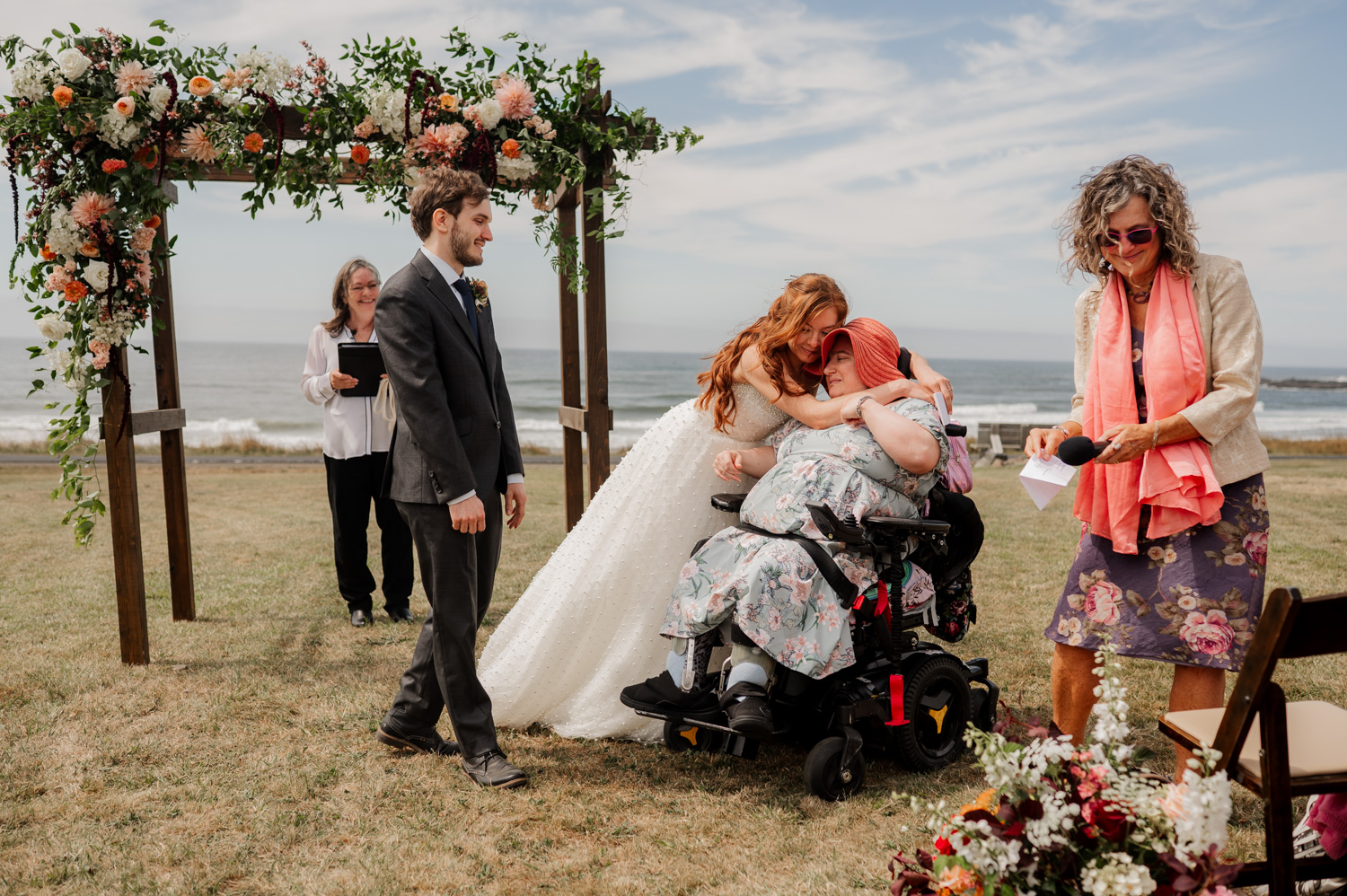 A bride embraces a person in a wheelchair at an outdoor wedding at the Yachats Inn, with several people standing nearby under a floral arch.