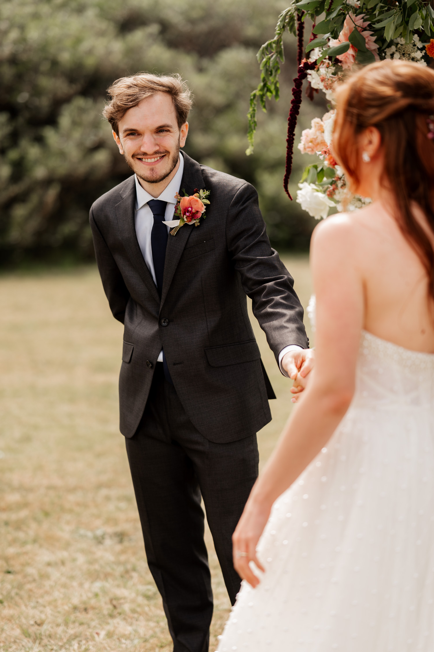 The groom in a dark suit smiles while holding hands with his bride in a white dress during their outdoor ceremony at the Yachats Inn.