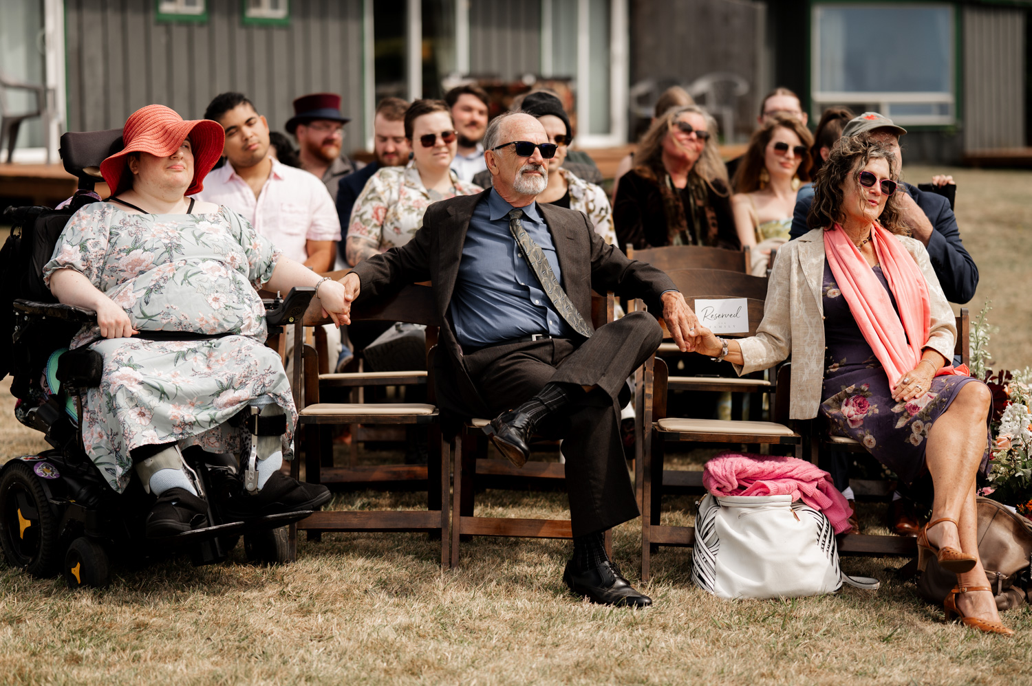 A group of people wearing formal attire are seated outdoors at the Yachats Inn, holding hands for wedding photos. One person in a wheelchair is part of the group.